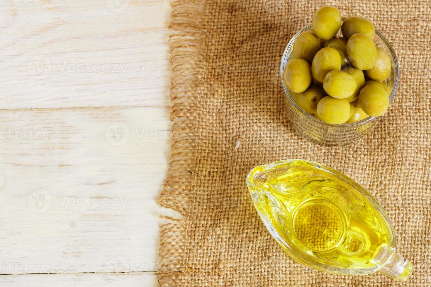 Top view of glass sauceboat with extra virgin olive oil and fresh green olives on burlap cloth on wooden table. photo