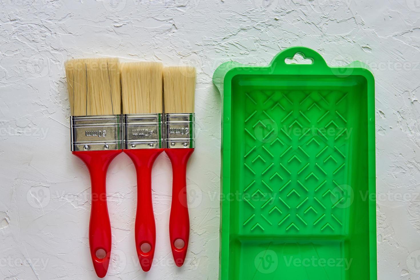 Three brushes with red handles and green paint tray on white concrete background. tools and accessories for home renovation. Top view photo