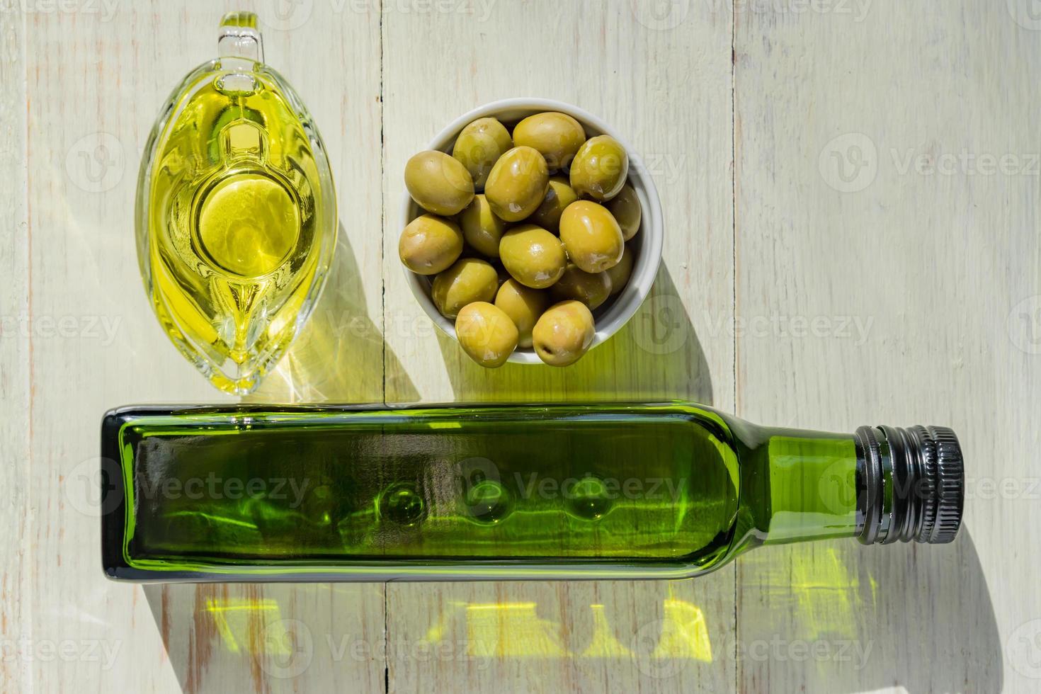 salsera de vidrio con aceite de oliva virgen extra, aceitunas verdes frescas y botella sobre mesa de madera. foto