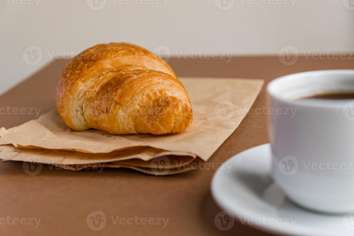 Tasty breackfast. French croissant served on craft paper and cup of black coffee or espresso on brown background. Copy space. photo