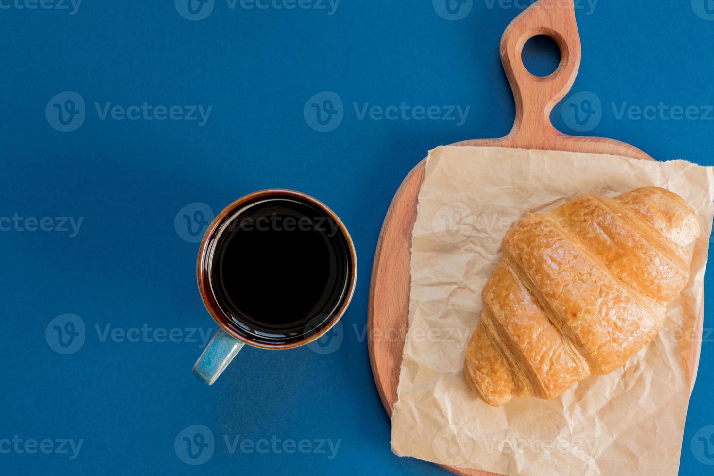 top view of cup of black coffe and croissant on a cutting board and craft paper on blue background with copy space. Morning breakfast in french style. photo