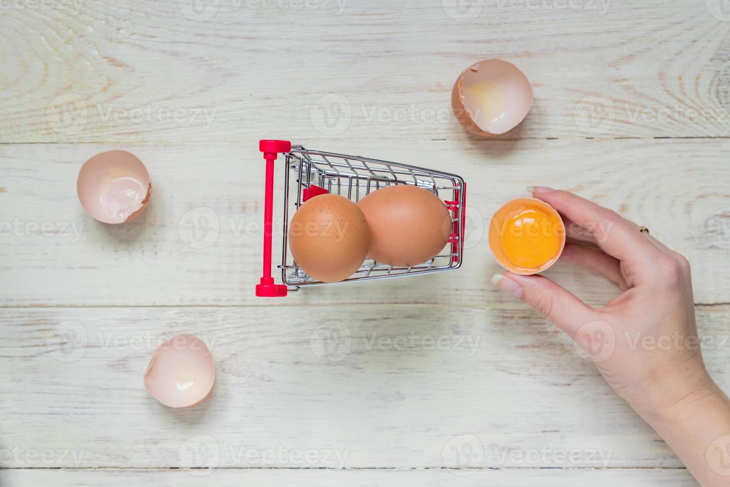 vista superior de huevos de pollo crudos marrones en carrito de compras y mano de mujer sosteniendo yema sobre fondo de madera foto