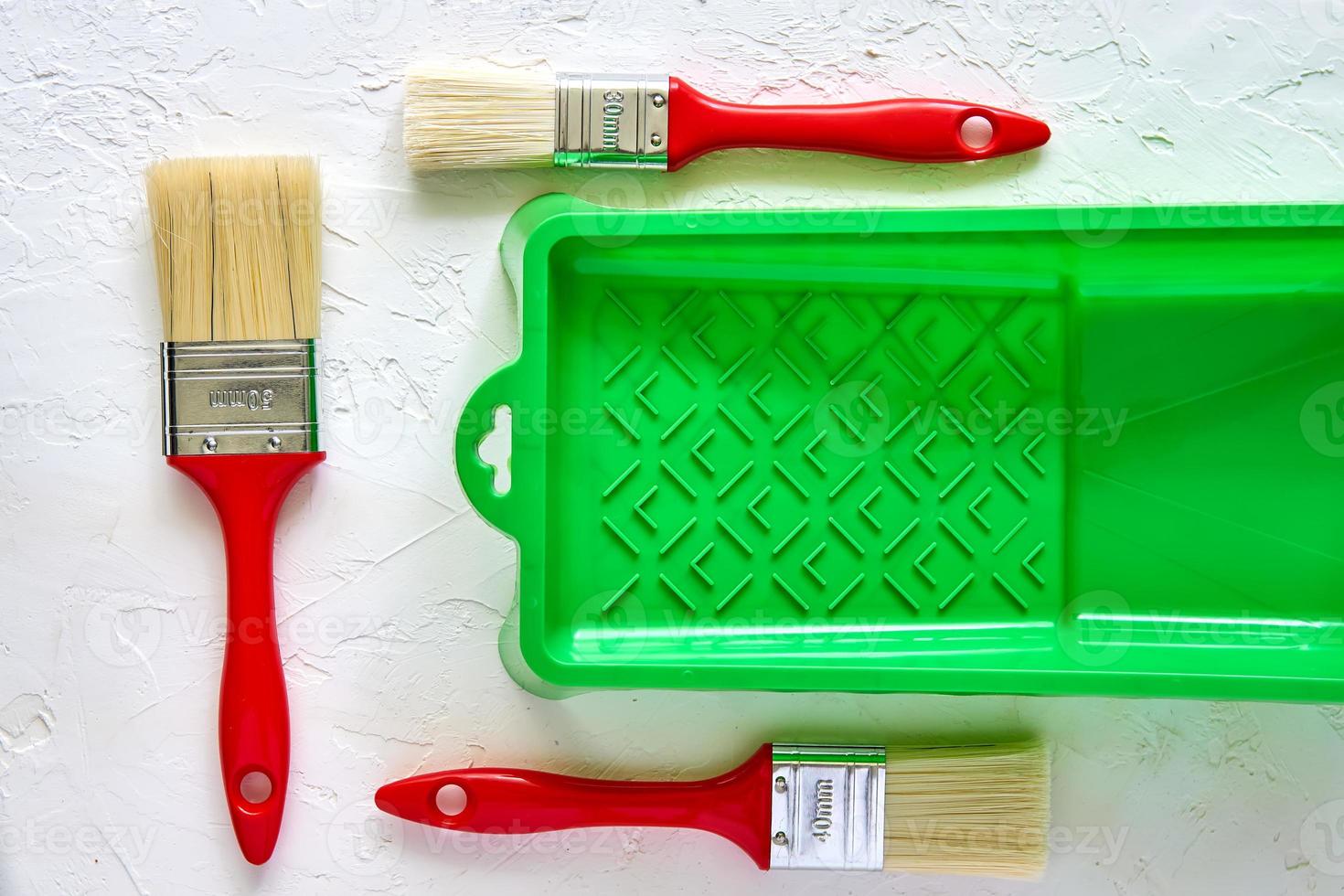 Three brushes with red handles and green paint tray on white concrete background. tools and accessories for home renovation. Top view photo