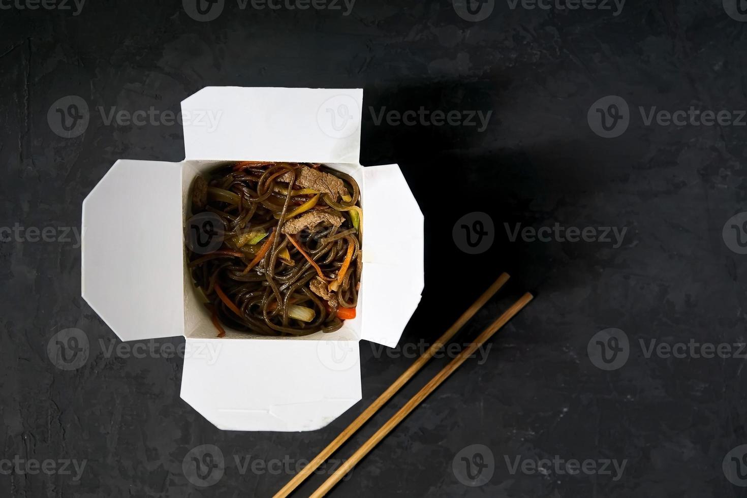 Asian restaurant food delivery. Soba noodles with meat, vegetables and soy sauce in white take-out paper box on black background with chopsticks, top view, copy space photo