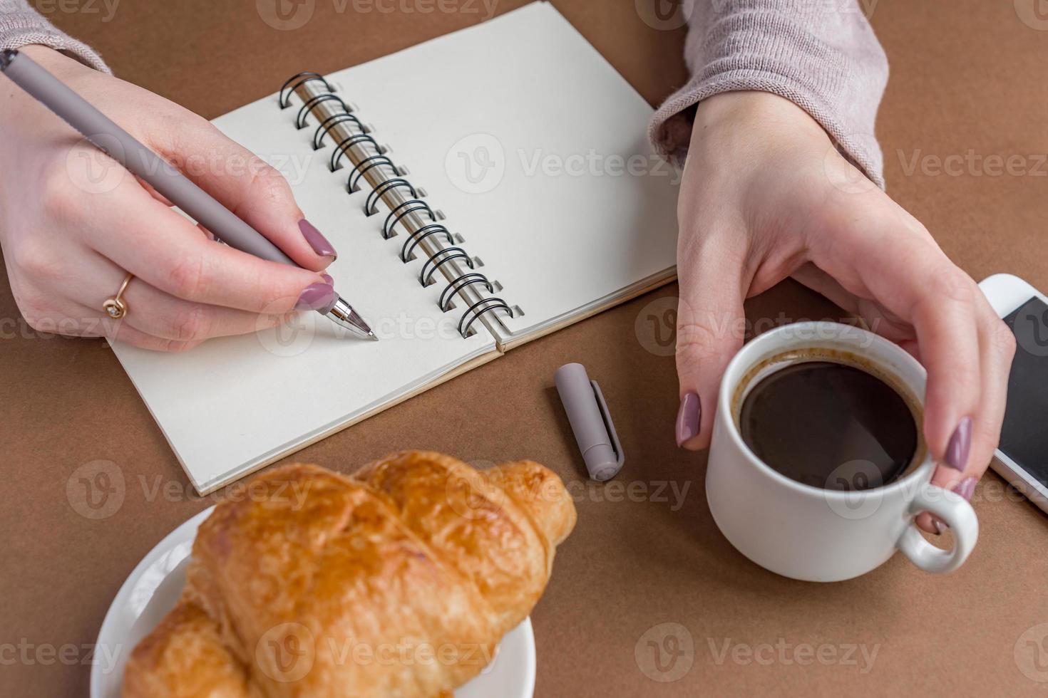 mujer con manicura escribiendo en el cuaderno. autónomo trabajando al aire libre. pausa para el café con croissant foto