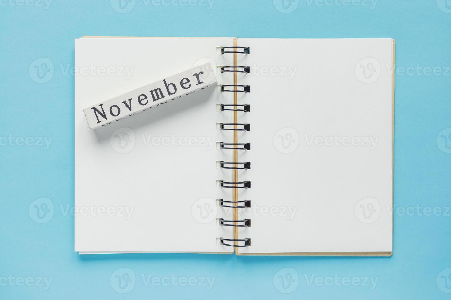 Clean spiral note book for notes and messages and november wooden calendar bar on blue background. Minimal business flat lay photo