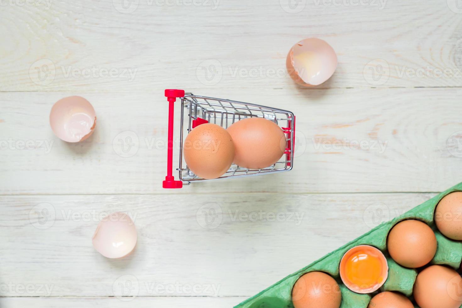 vista superior de huevos de pollo crudos marrones en caja de cartón de huevo verde y carrito de compras sobre fondo de madera blanca foto