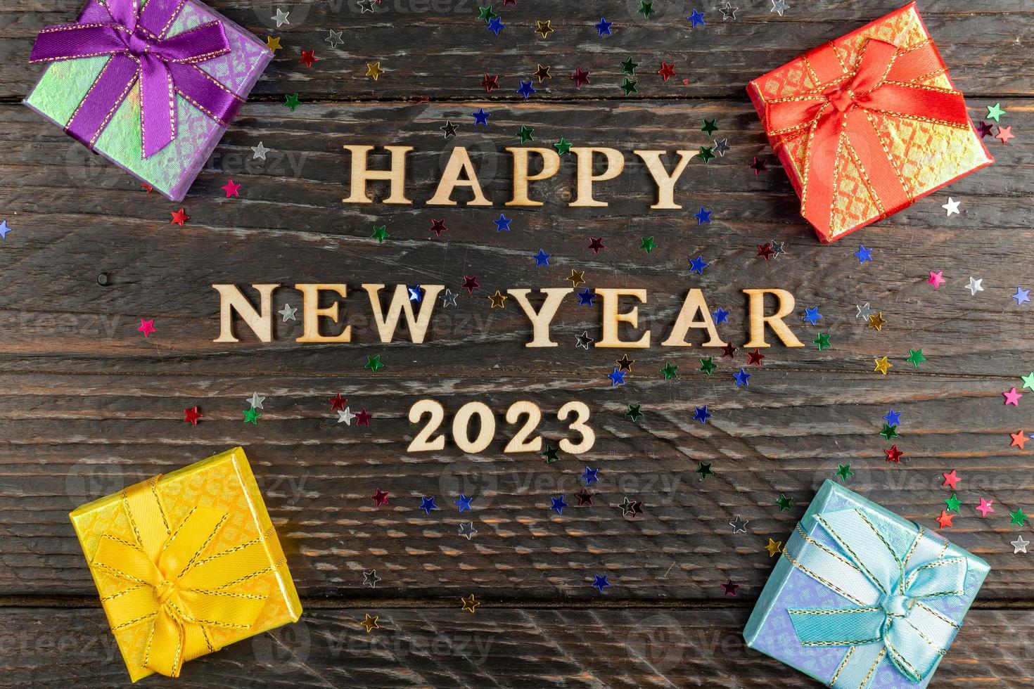 Multicolored gift boxes with tied bows. New year presents on wooden table with scattered confetti stars. Text Happy new year 2023. Festive greeting card. Flat lay. photo