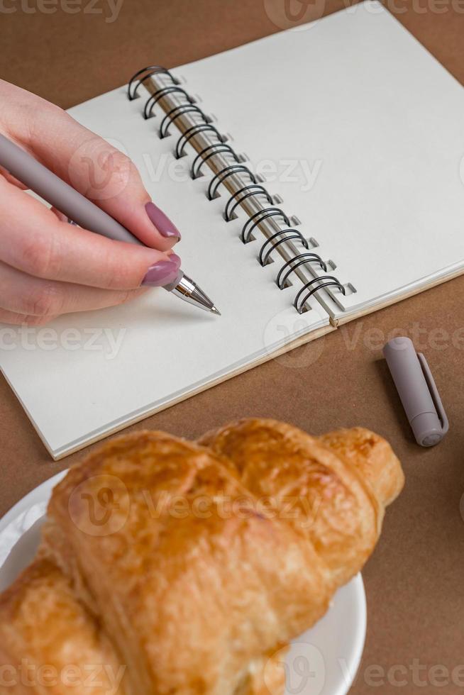 mujer con manicura escribiendo en el cuaderno. autónomo trabajando al aire libre. sabroso croissant en un plato blanco sobre la mesa. foto