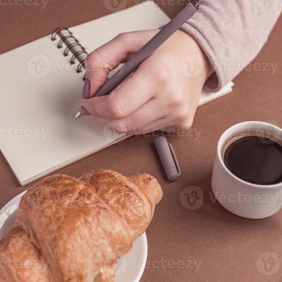 mujer mano izquierda con pluma escribiendo en el cuaderno en la cafetería. freelancer trabajando al aire libre. coffe break con croissant y espresso foto