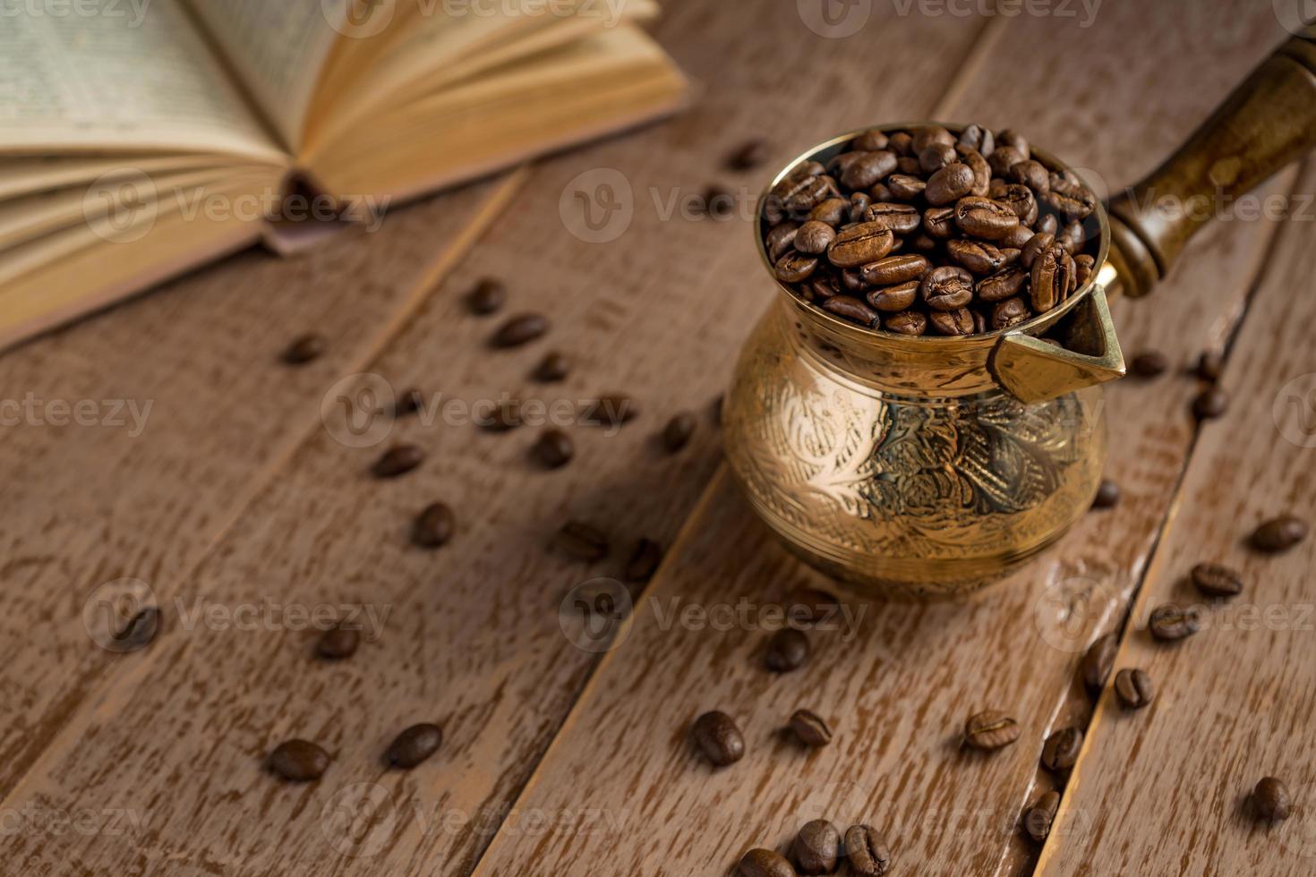 granos de café recién tostados en cezve, cafetera tradicional turca, libro abierto y taza sobre una mesa de madera. foto