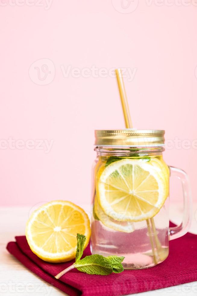 Citrus lemonade water with sliced lemon and mint, healthy and detox water drink in summer on wooden table with pastel pink background photo