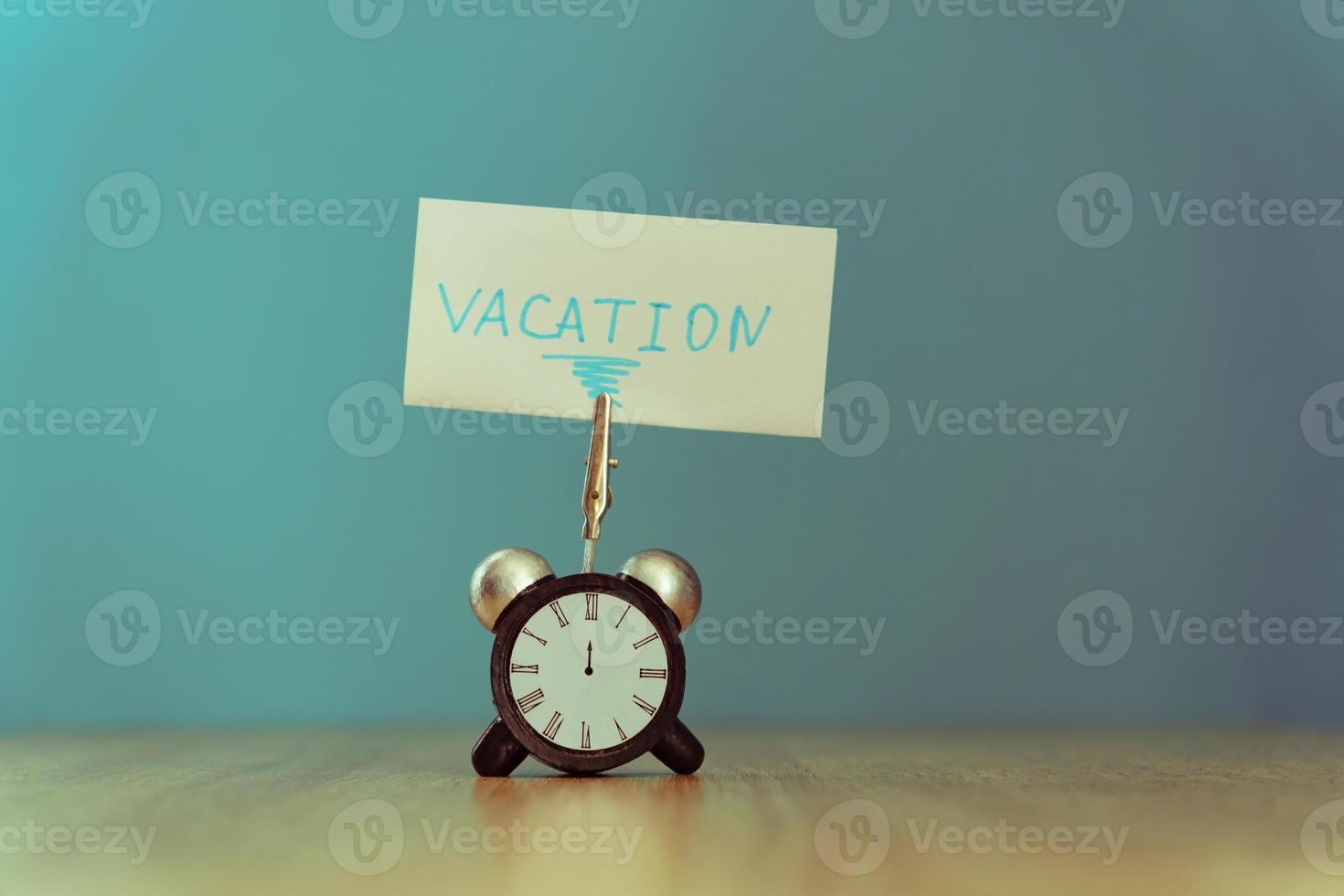 Alarm clock and banner with handwritten phrase vacation on wooden table. Vacation concept. Toned photo