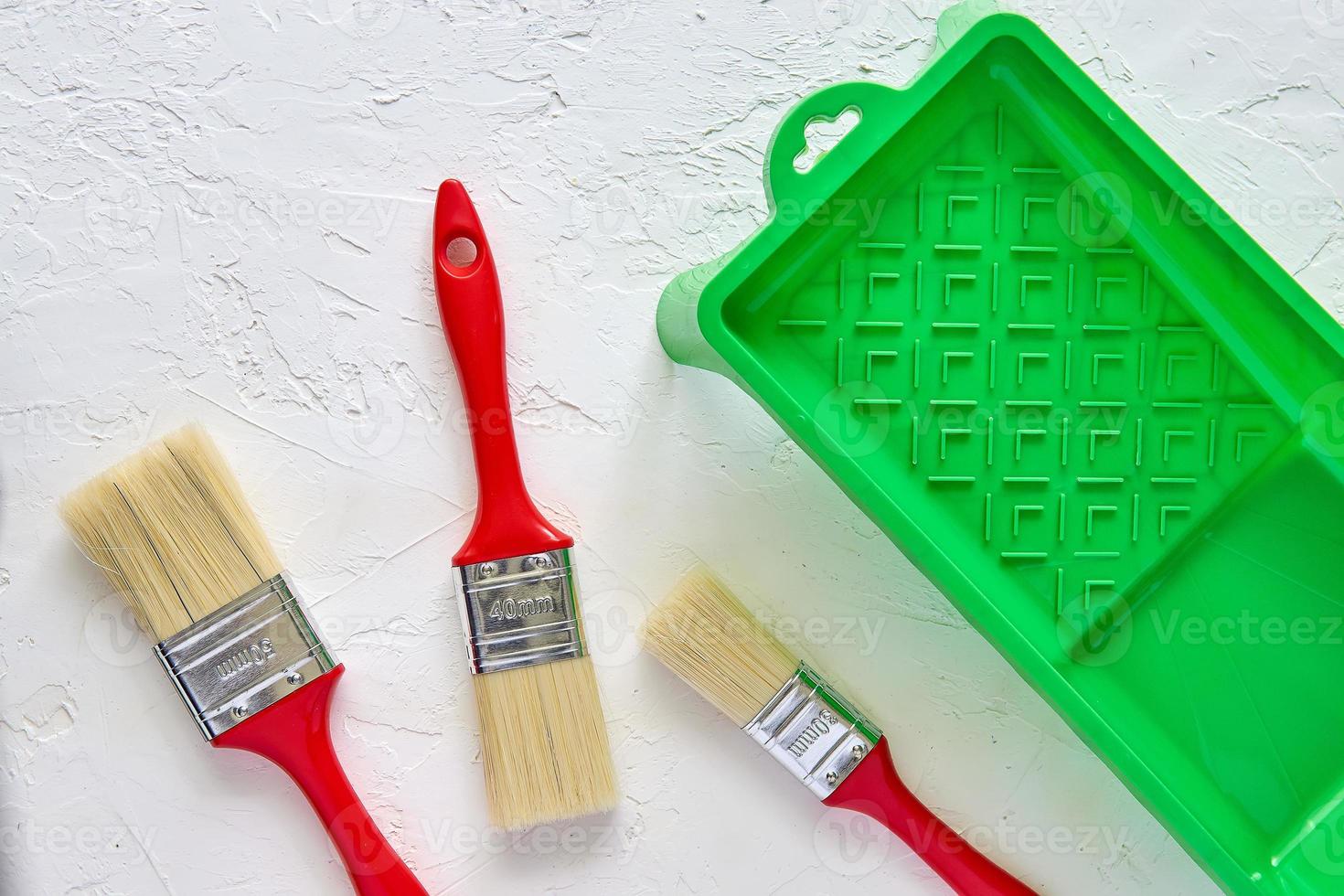 tres cepillos con asas rojas y bandeja de pintura verde sobre fondo de hormigón blanco. herramientas y accesorios para la renovación del hogar. vista superior foto