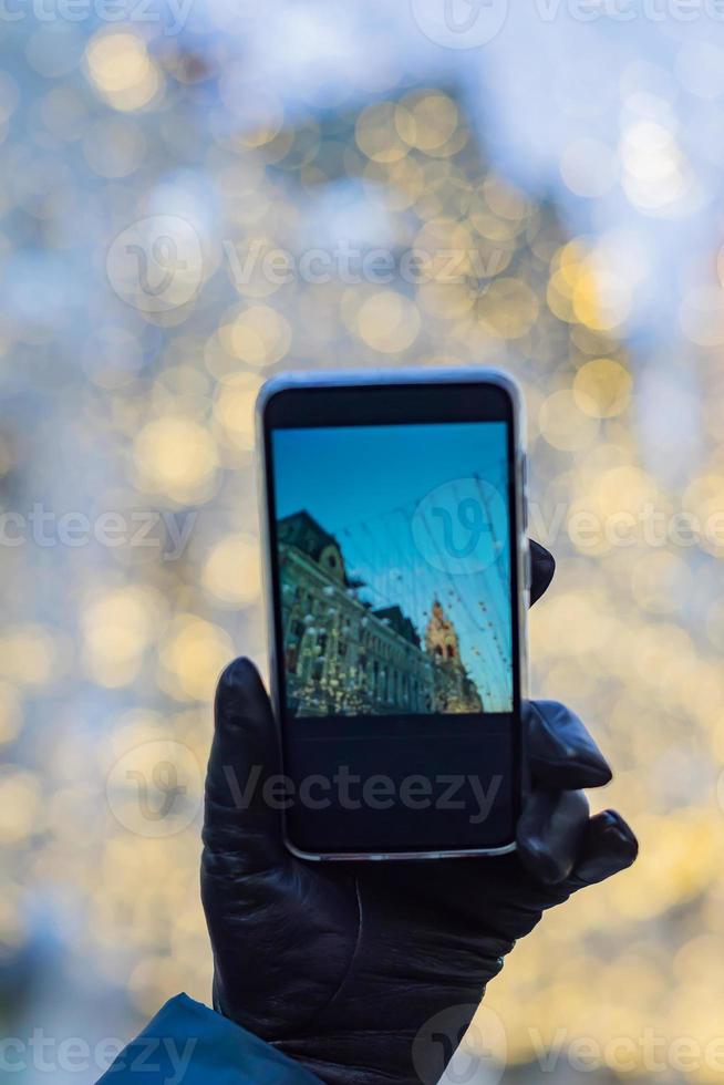 mano de mujer en un guante de cuero negro con un smartphone negro para tomar una foto de la ciudad de Navidad en un soleado día de invierno. fondo bokeh