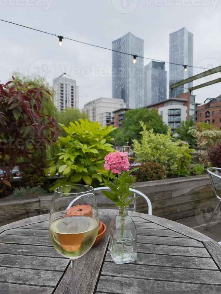 Copa de vino blanco junto a una flor en una mesa de un pub al aire libre con rascacielos en la distancia en Castlefield en Manchester, Reino Unido foto