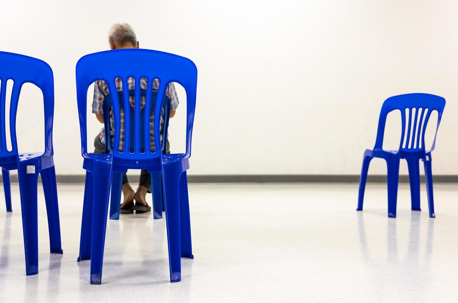 A view from behind a blue plastic chair that is distanced from a single elderly man. photo