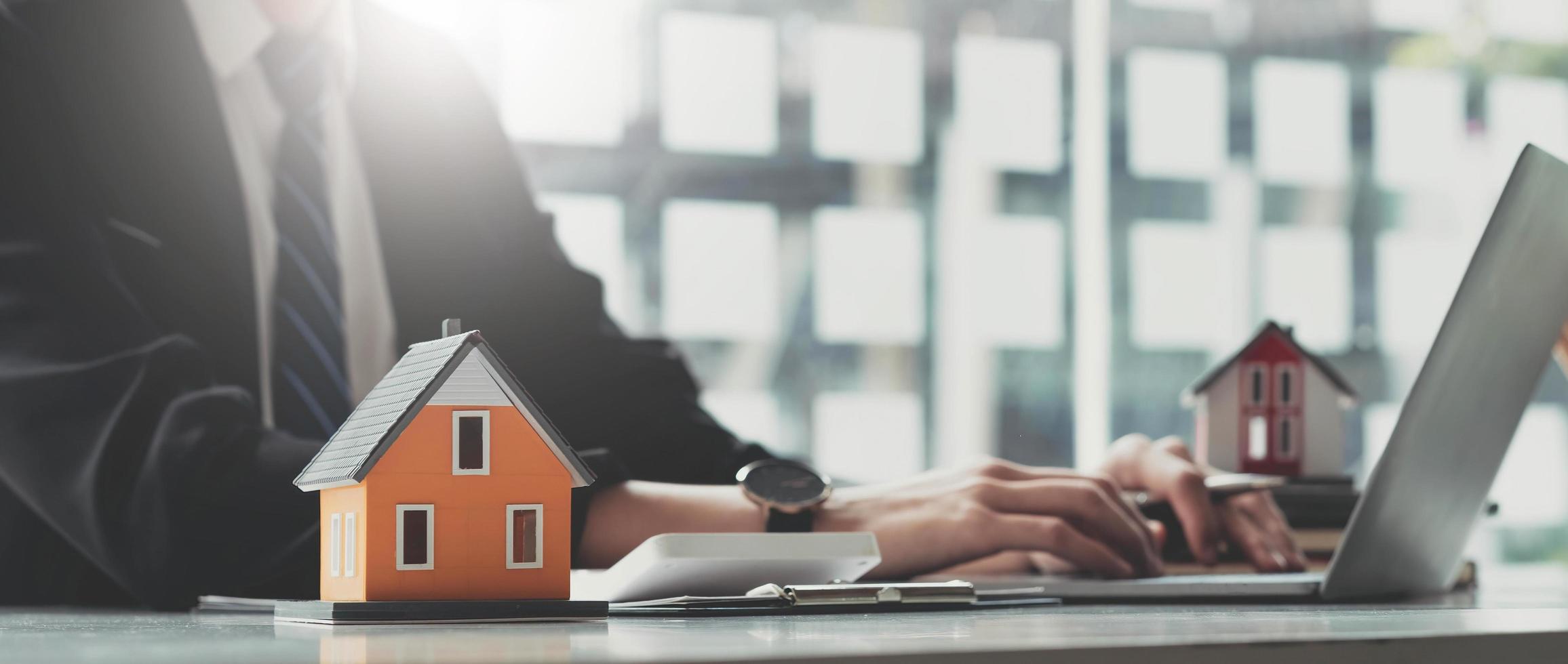 Photo of a house model putting on a clipboard at the wooden working desk over a real estate using a computer laptop as a background.