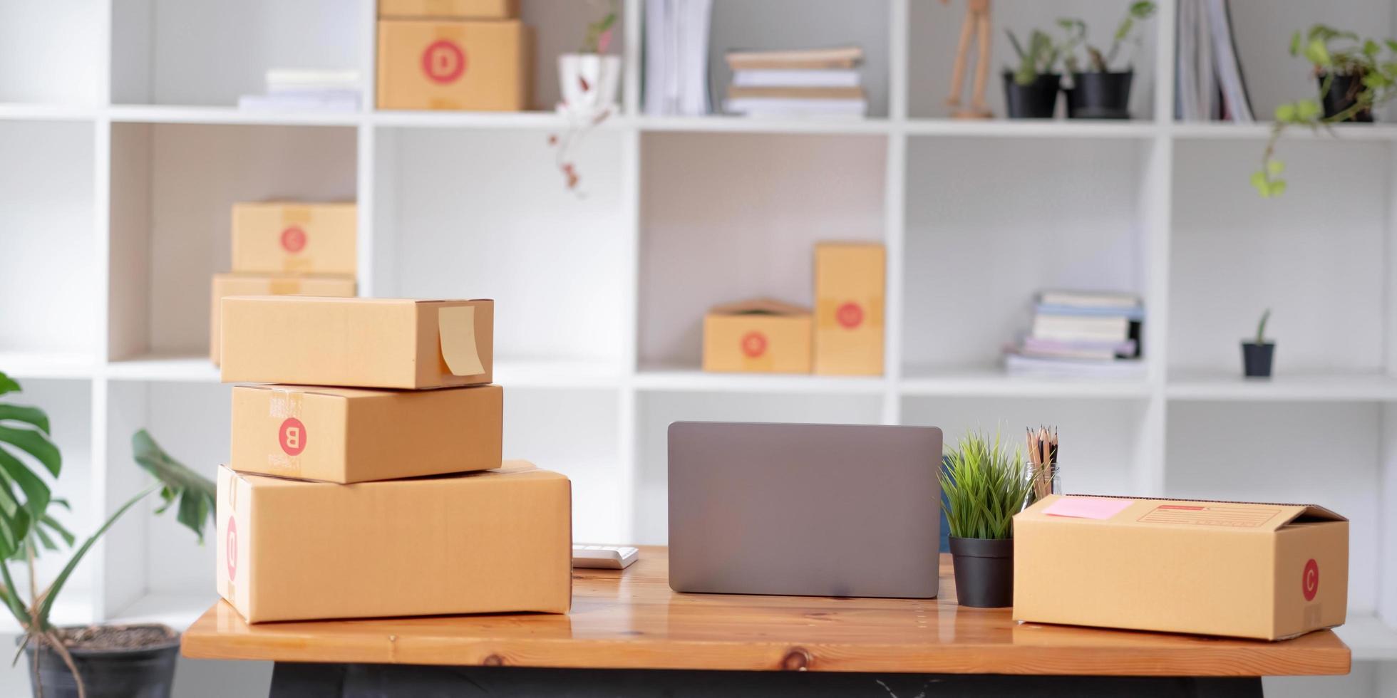 cajas de paquetes en el estante y bolsas de compras de color colocadas cerca de la computadora portátil en la mesa. Las pequeñas empresas que compran en línea en el embalaje de la oficina en casa en el fondo son negocios populares. foto