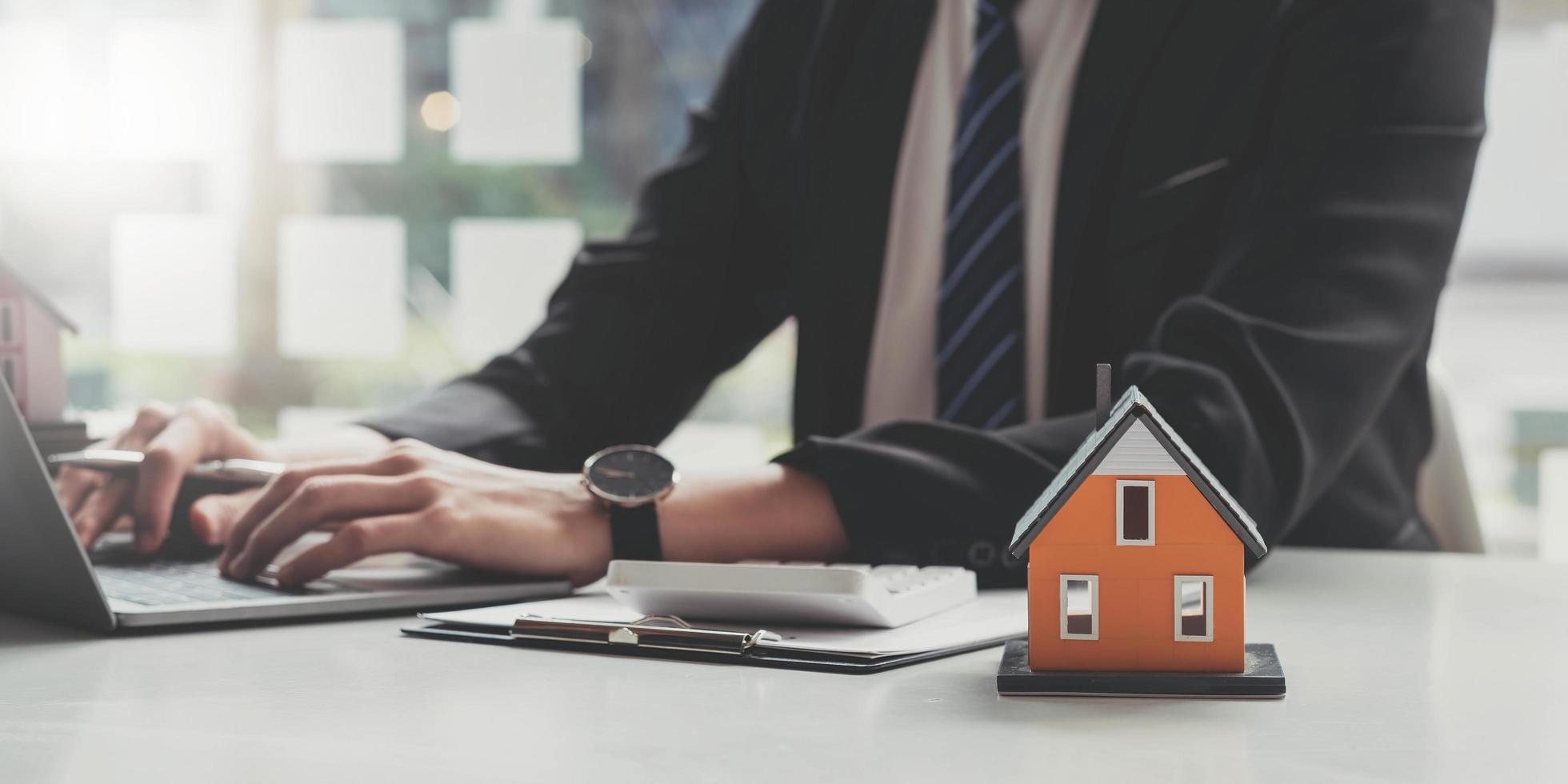 Photo of a house model putting on a clipboard at the wooden working desk over a real estate using a computer laptop as a background.