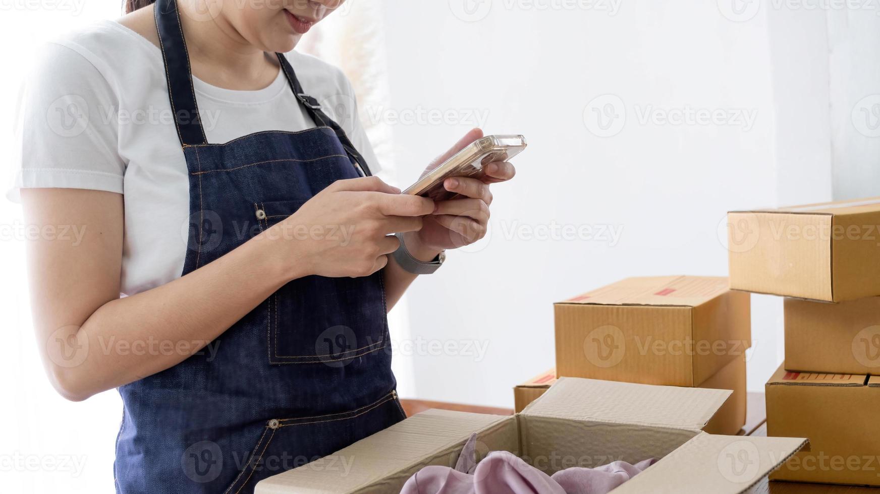 Hands of Asian woman entrepreneur holding smartphone with cardboard boxes, computer laptop, online selling equipments. Packing, business and technology concept. Top view, copy space photo