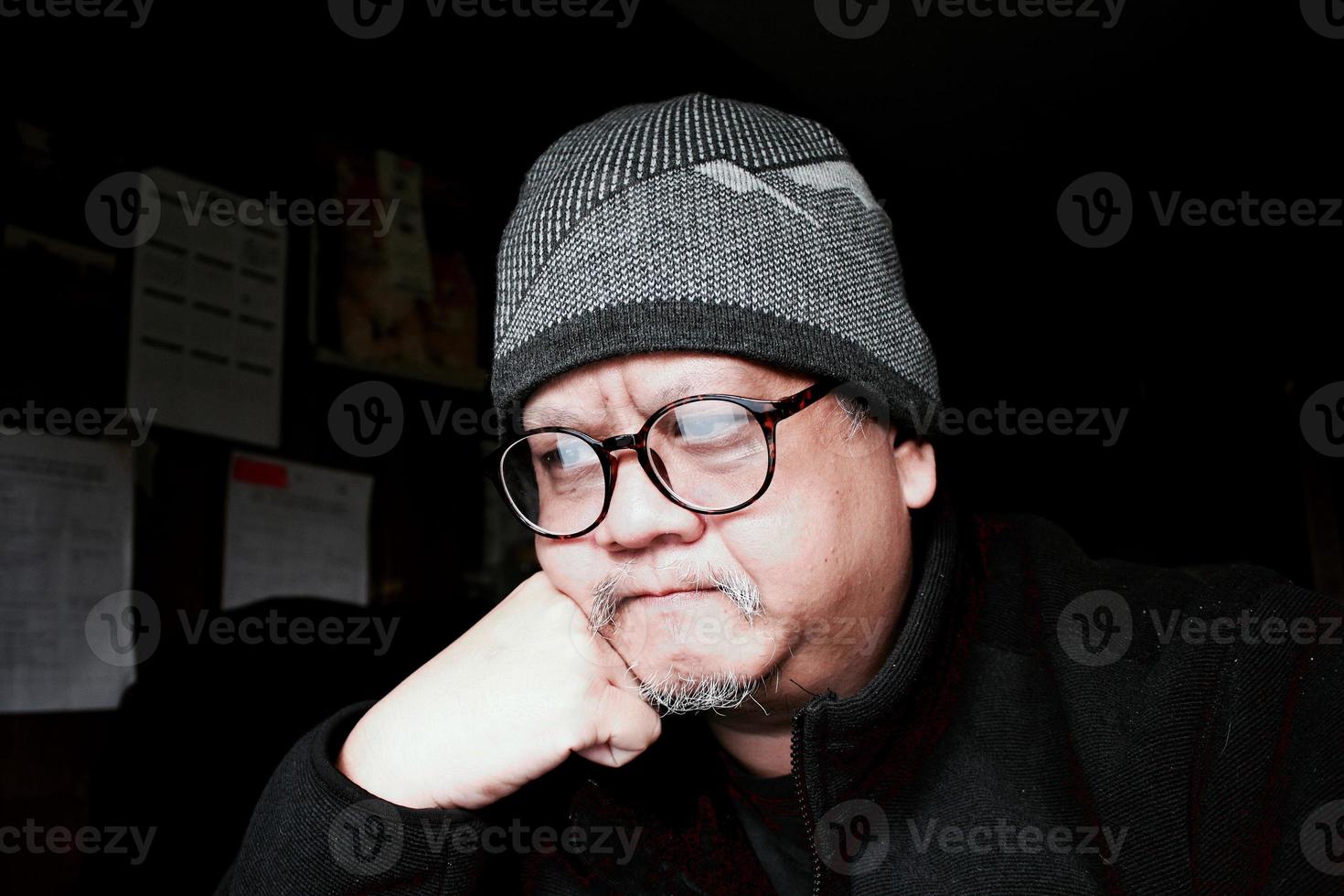 hombre asiático barbudo con chaqueta negra y gorro de punto. trabajando en una habitación oscura y poniendo sus manos en su barbilla foto