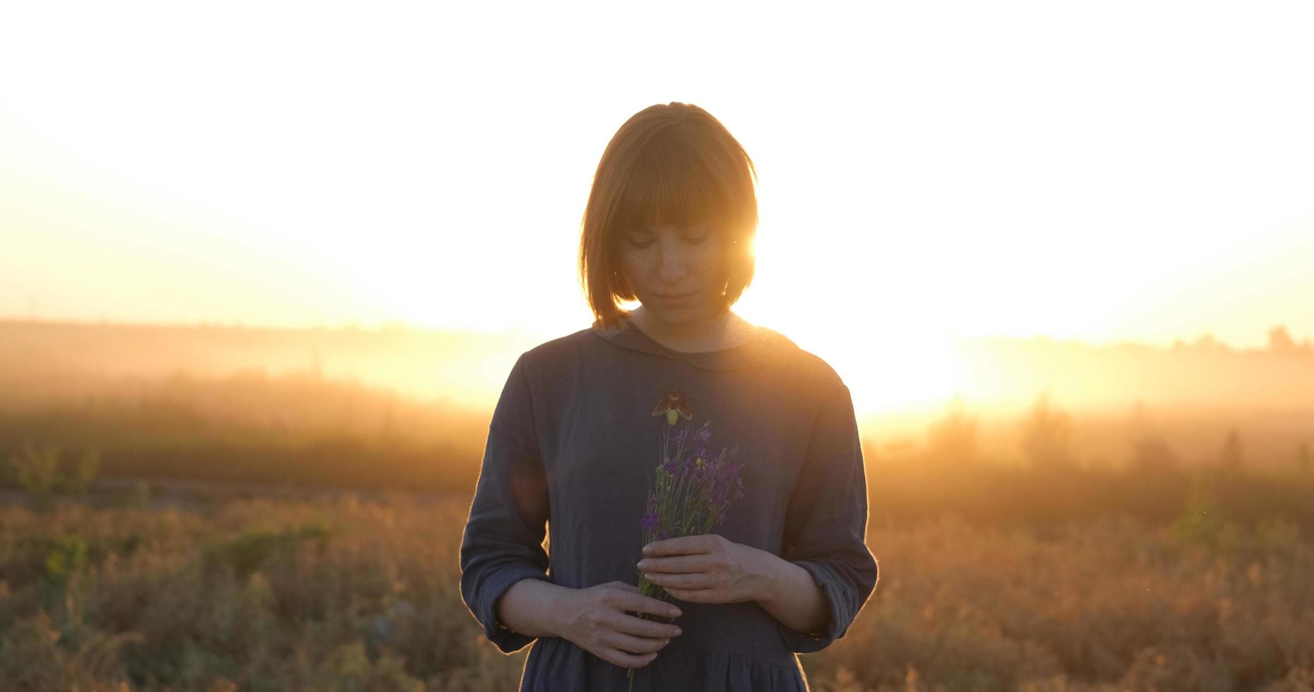 joven pelirroja con hermoso vestido boho relajándose en el campo durante la puesta de sol nublada, mujer al aire libre con ramo en las manos foto