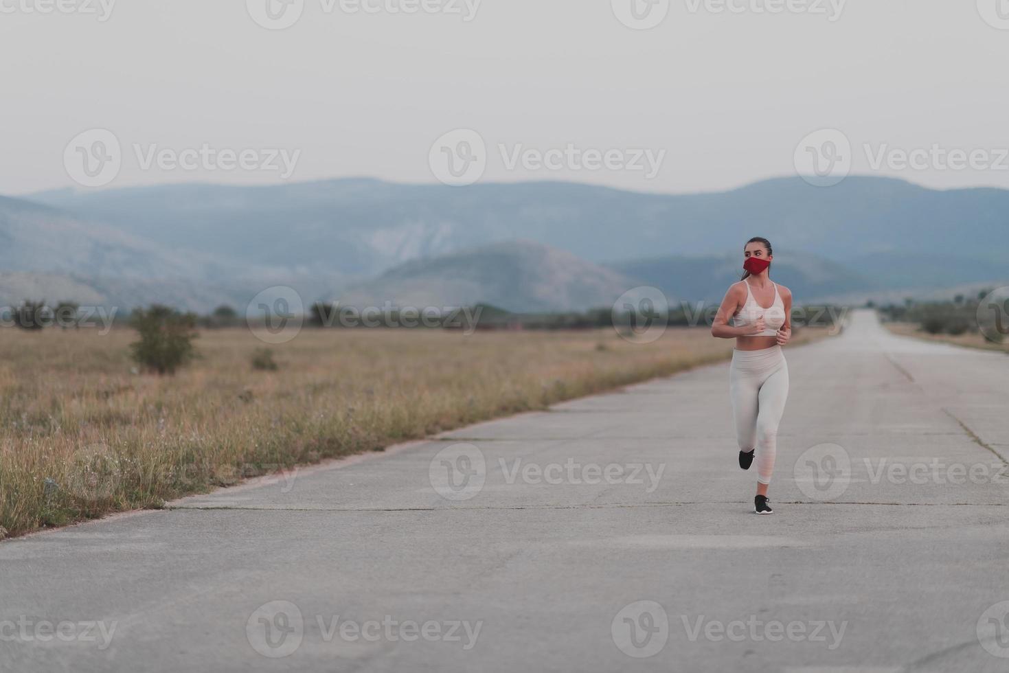 Determined fitness woman in short clothes wearing red protective face mask running outdoors in the city during coronavirus outbreak. Covid 19 and physical jogging activity sport and fitness. photo
