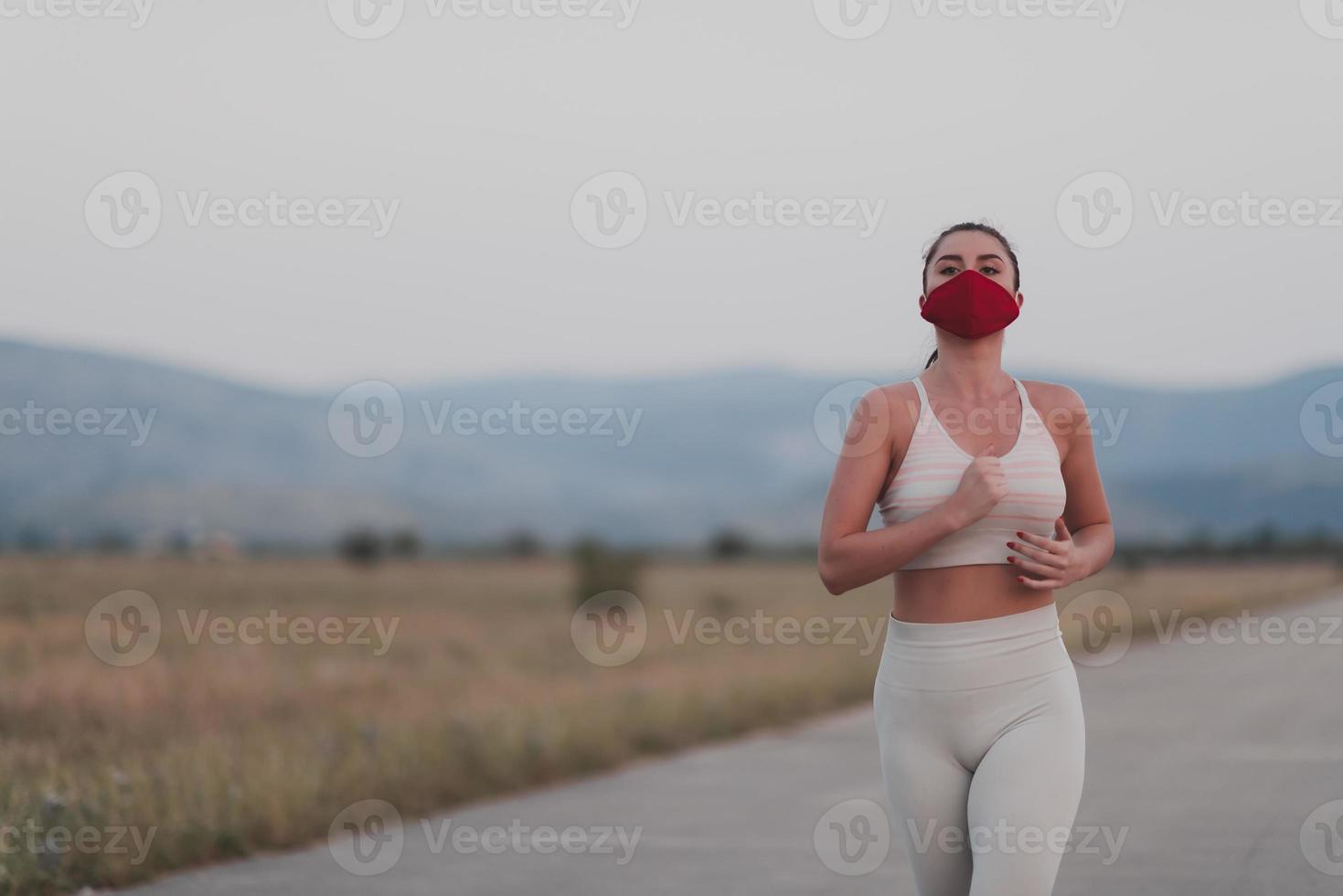 Determined fitness woman in short clothes wearing red protective face mask running outdoors in the city during coronavirus outbreak. Covid 19 and physical jogging activity sport and fitness. photo