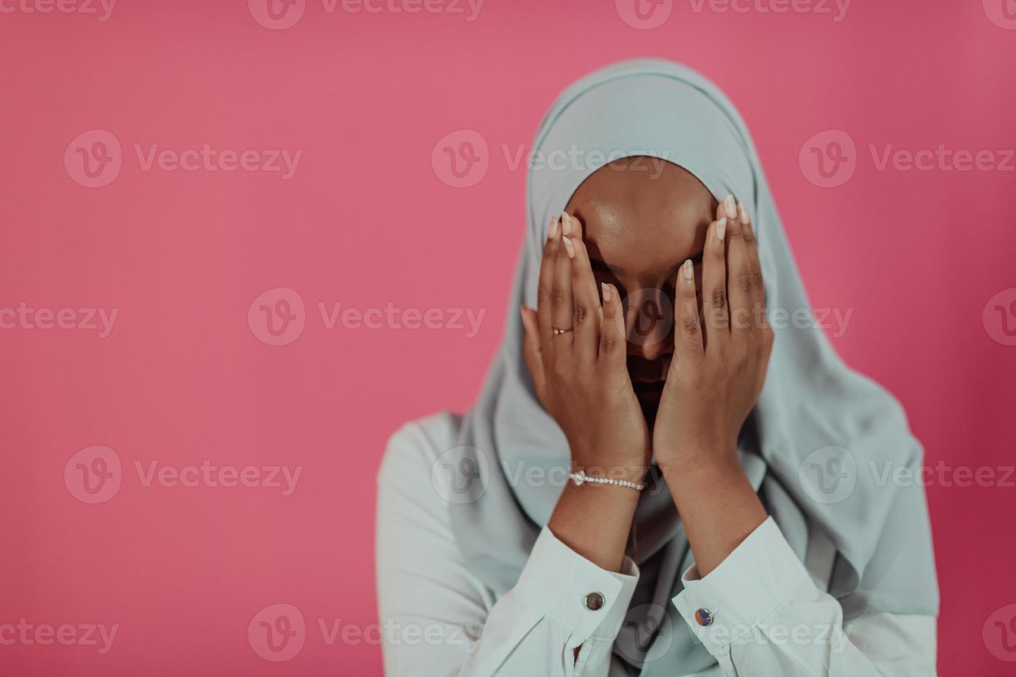 Modern African Muslim woman makes traditional prayer to God, keeps hands in praying gesture, wears traditional white clothes, has serious facial expression, isolated over plastic pink background photo