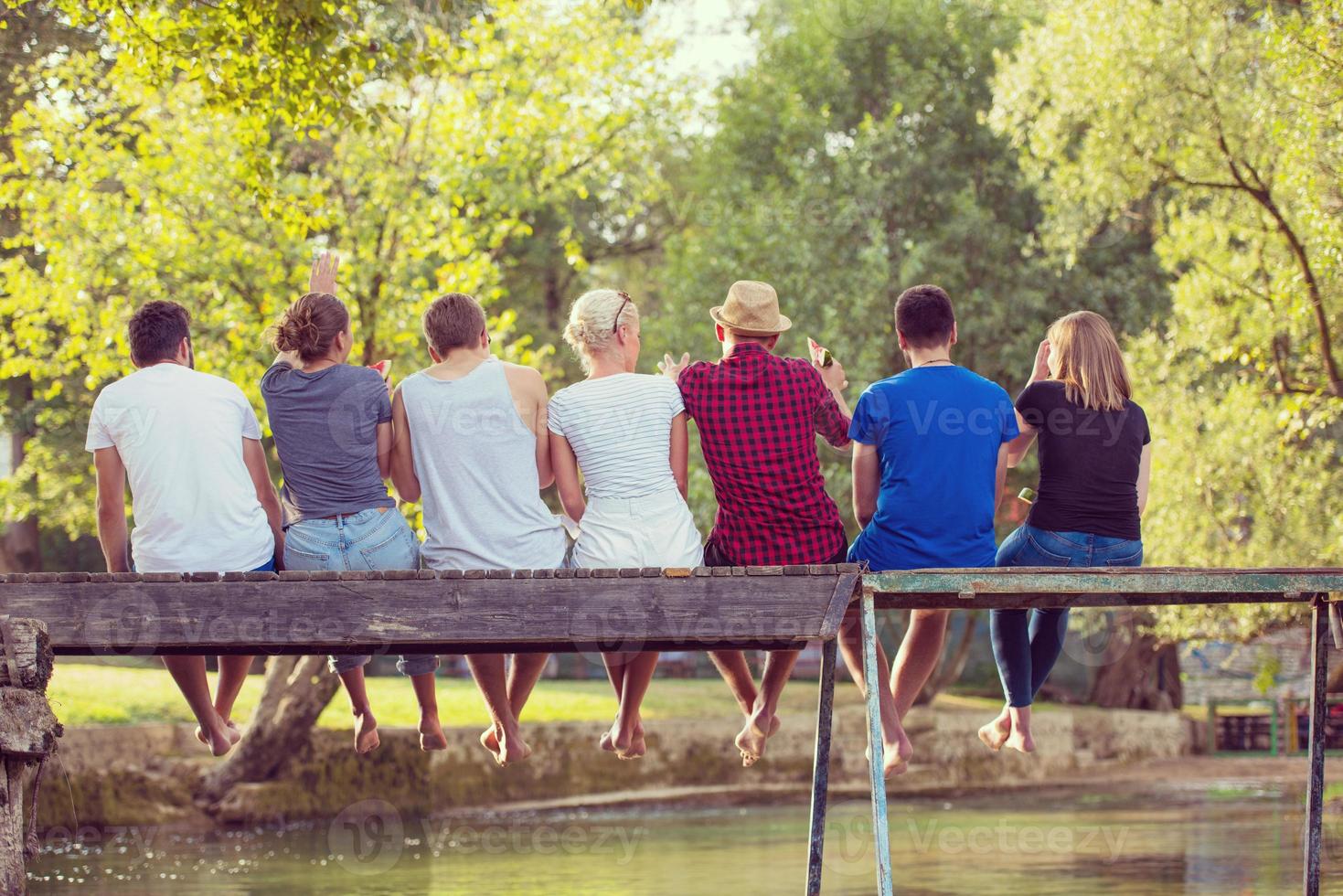 rear view of friends enjoying watermelon while sitting on the wooden bridge photo