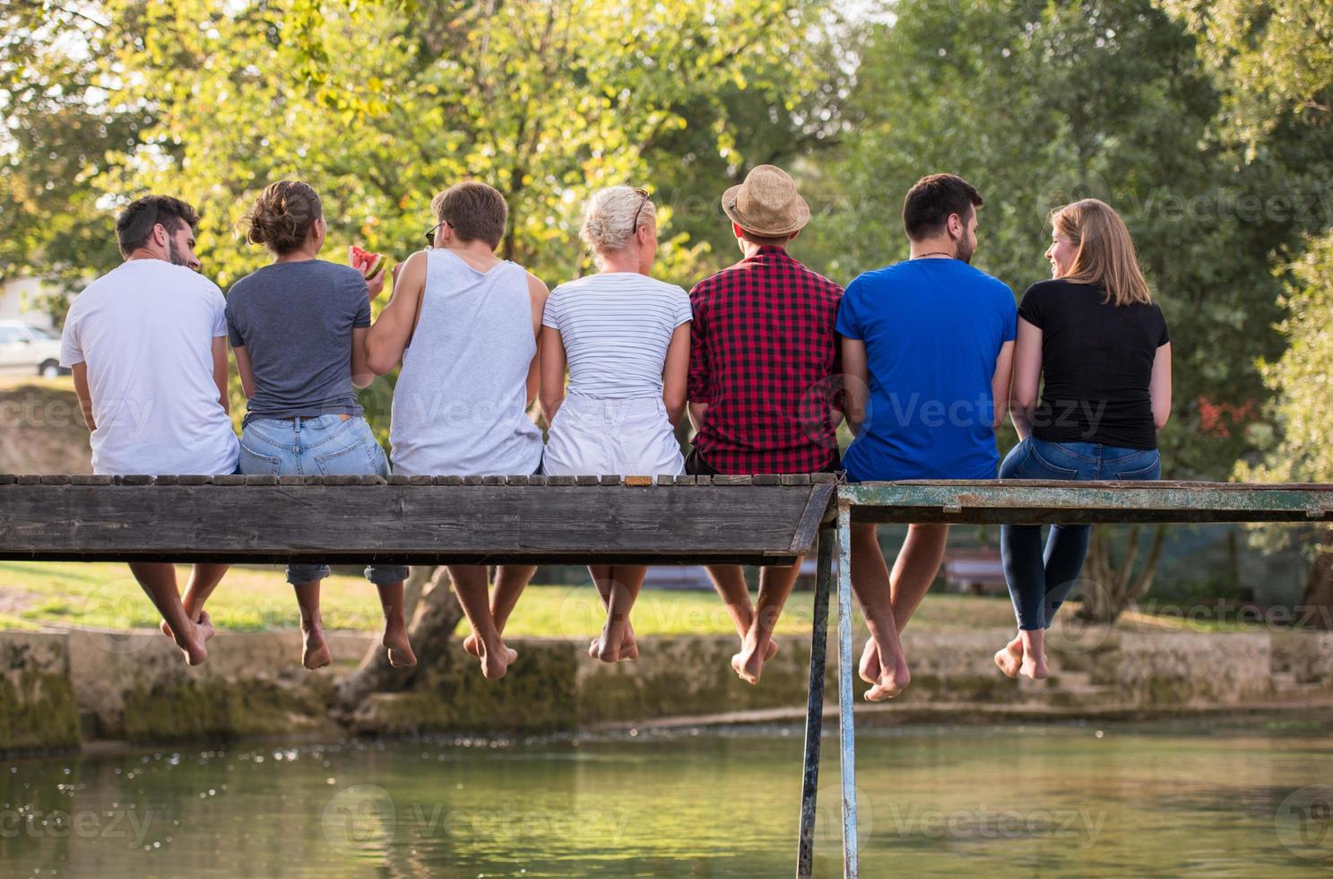 rear view of friends enjoying watermelon while sitting on the wooden bridge photo