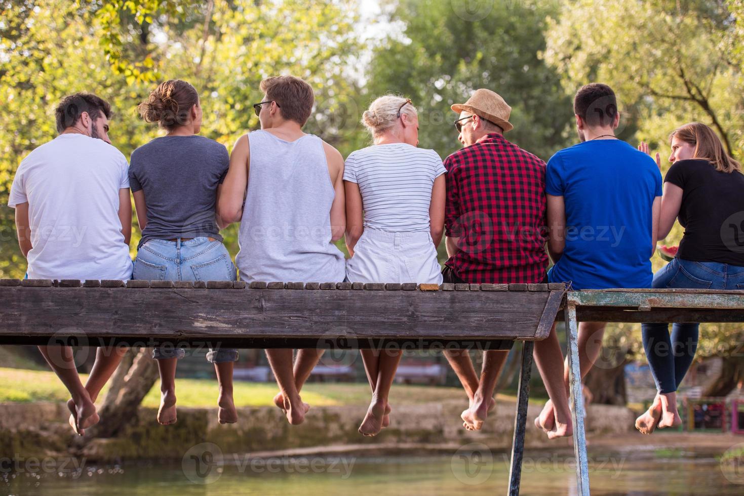 rear view of friends enjoying watermelon while sitting on the wooden bridge photo
