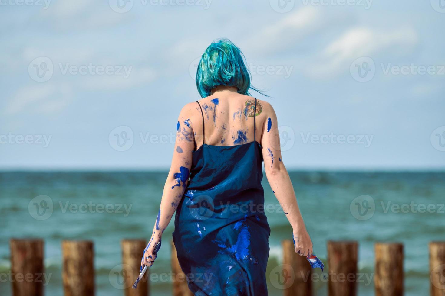 Artistic blue-haired woman performance artist smeared with blue gouache paints dancing on beach photo