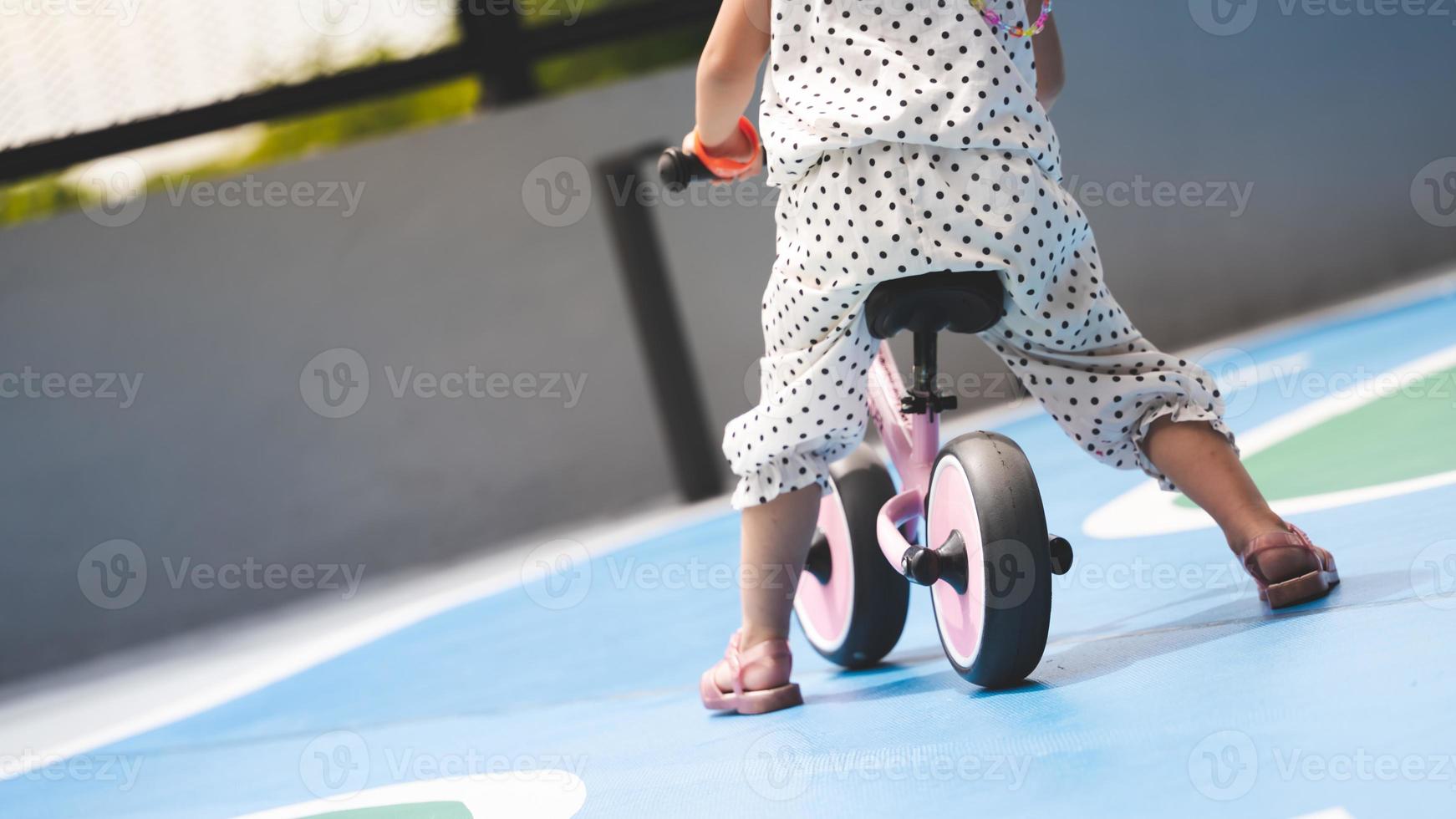 Toddler on bicycle, leg of small child sitting on bike, girl riding pink plow bike on playground road, learning to ride through playing concept. Empty space for entering text. Summer or spring time. photo