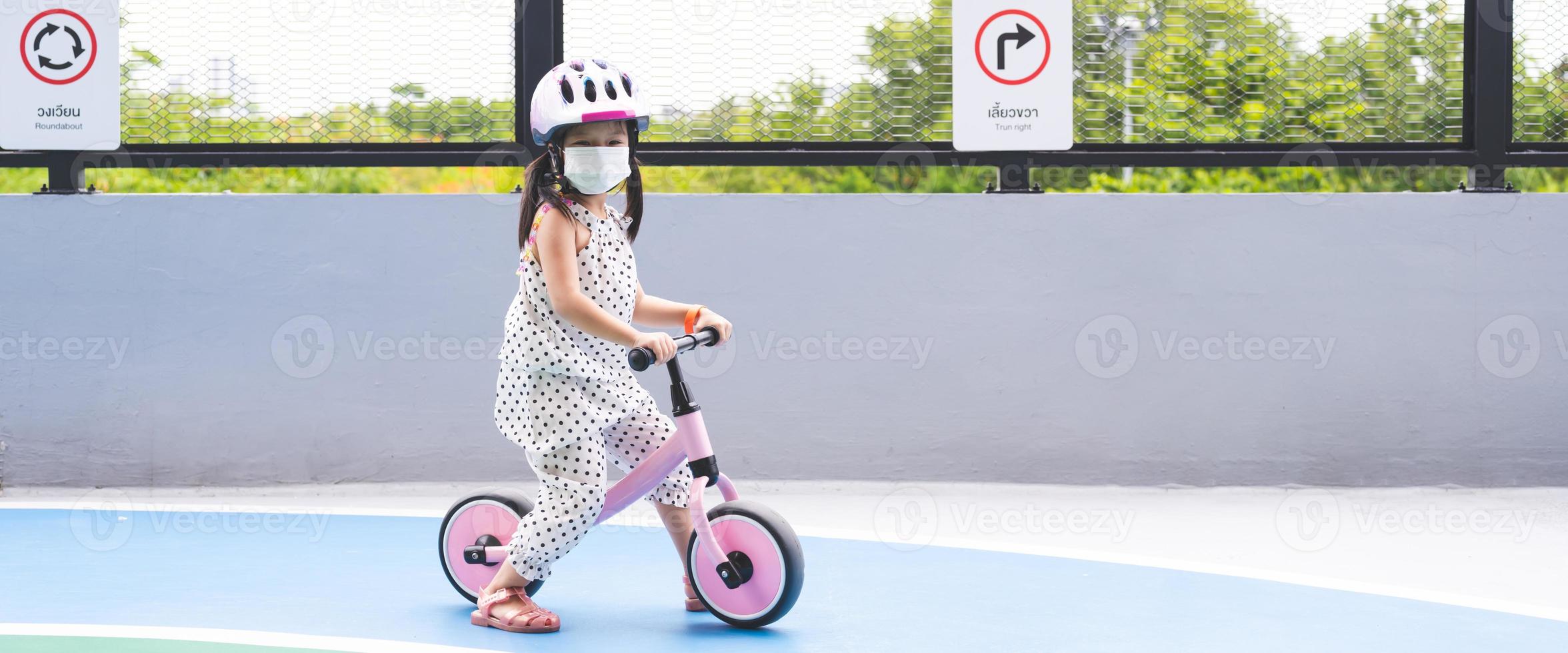 Cute Asian girl is playing on bicycle with her legs plowed on ground. Children wear helmets while practicing. Behind kid is traffic sign written in Thai and English that says Roundabout and Turn right photo