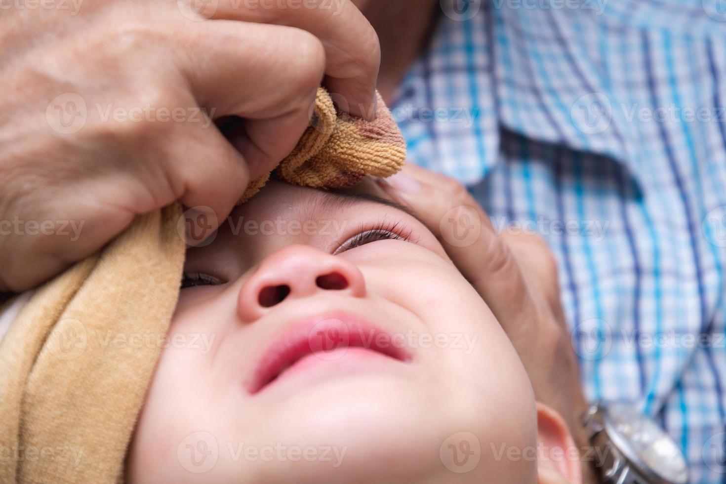 Boy is crying out of pain. Child bumps the head against solid. Baby is bulging. Adult is wrapping ice cube in cold compress on child. Closeup, crying son face from injury caused by home accident. photo