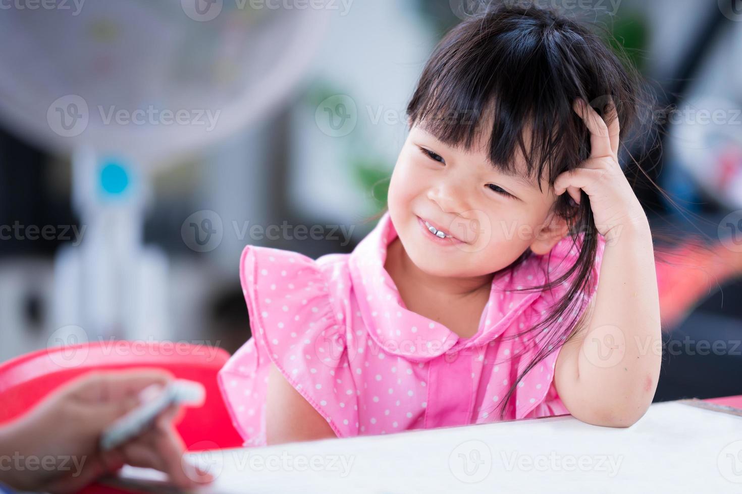 Adorable kid girl sweet smile in the small classroom at home school. Child thinking about concept her artwork with teacher. Children wearing pink shirt and polka dots pattern. Lady 4 years old. photo