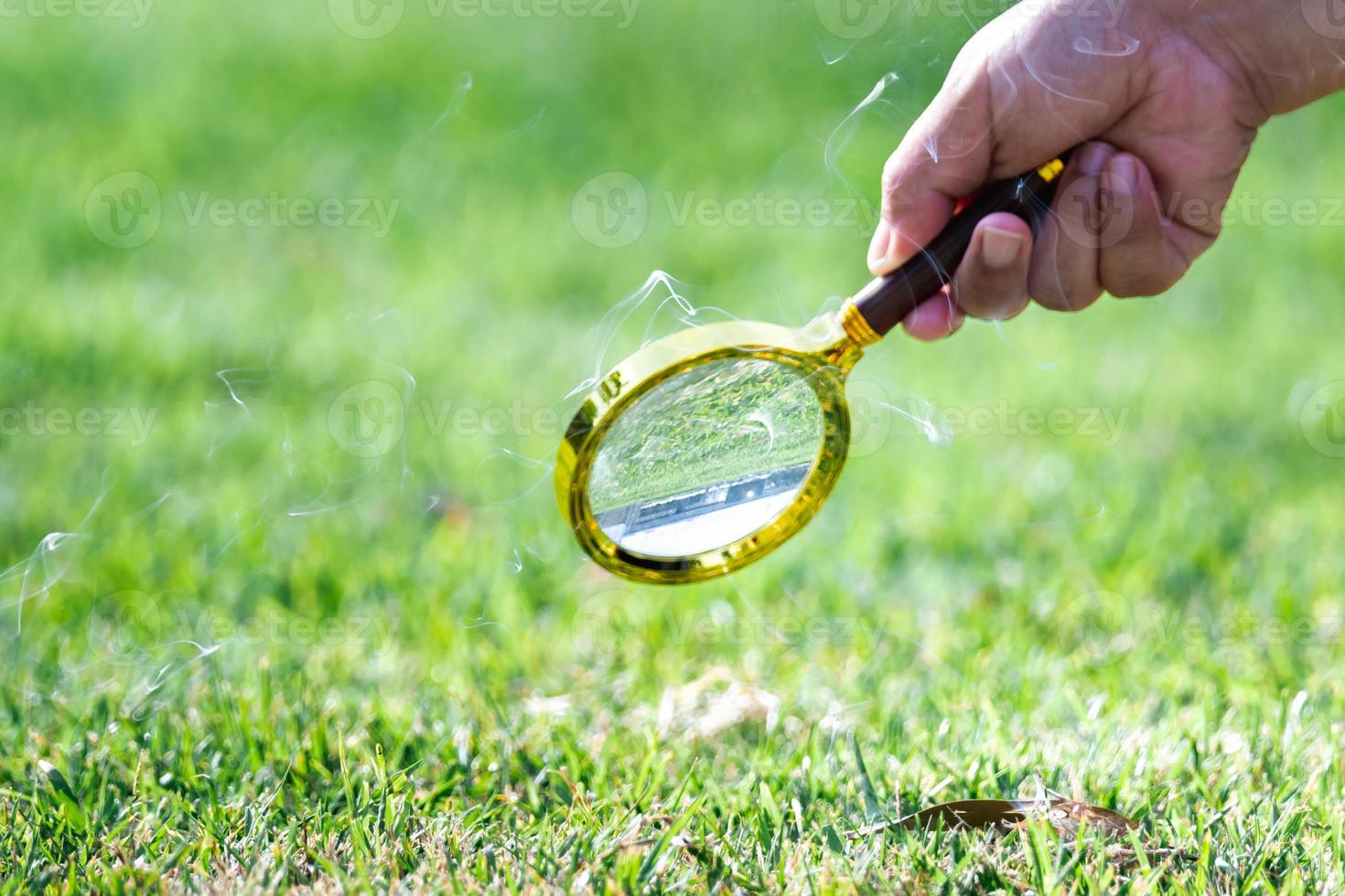 A human's hand holding a magnifying glass with golden edges is gathering sunlight from the sun onto the dry leaves on the green lawn. Smoke produced by burning leaves. photo