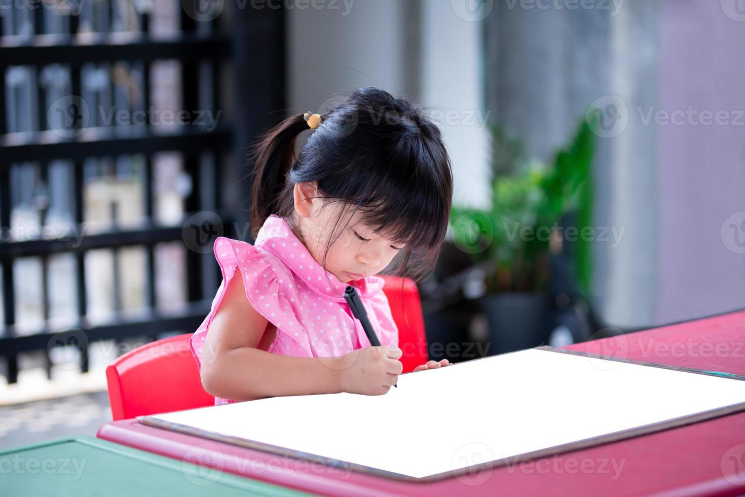 Cute Asian girl sitting on a drawing of myself on piece of paper that is pinned on plywood sheet. Child do art. Kid is cutting a line with black magic pen. Children 4 years is doing imaginative crafts photo