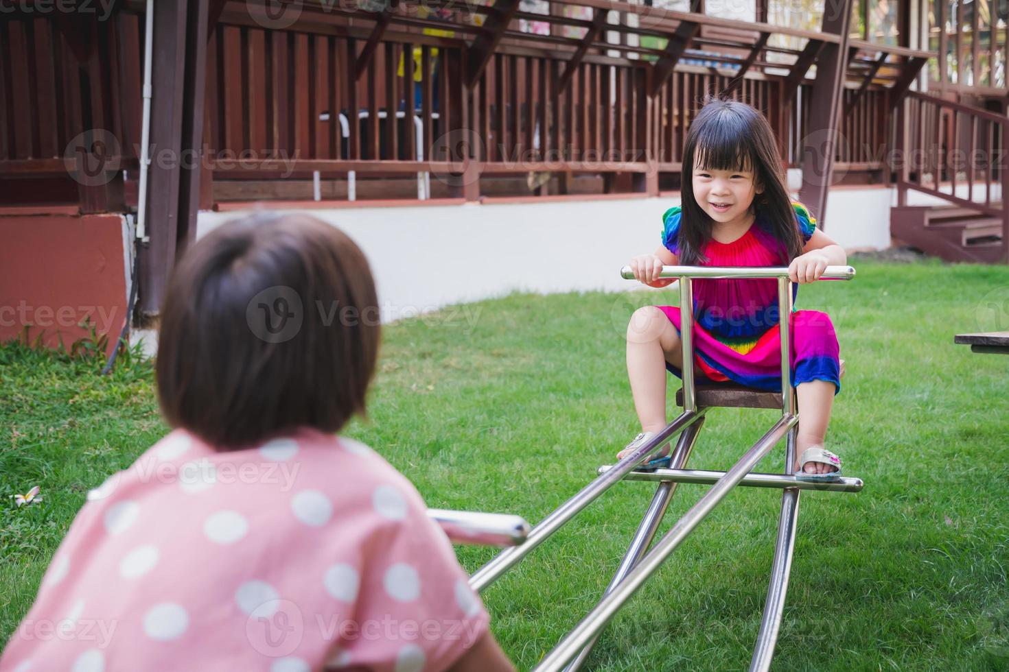 Two girl siblings playing teeter totter rocking with joy. Asian child have sweet smile. Children laughed brightly. 4-6 year old kid wearing colorful clothes. Play in the playground . Summer or spring. photo