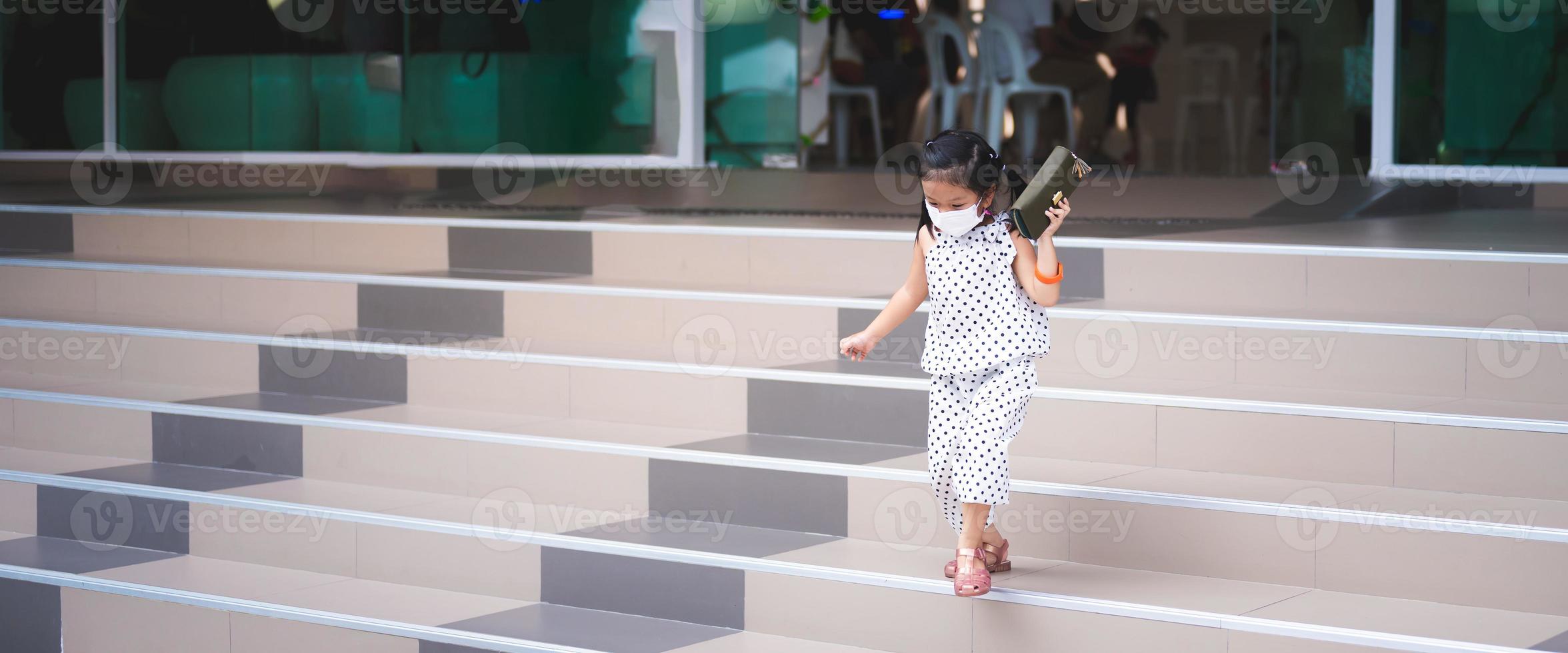 Portrait image child 5-6 years old. Little girl wearing white face mask for prevent toxic dust pm2.5 and virus. Cute kid was cautiously running down the stairs, holding purse in her hand. Empty space. photo