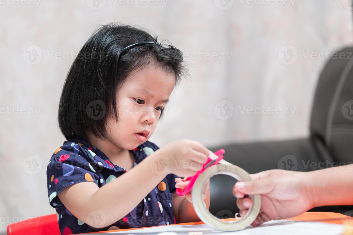 Asian little girl cutting paper tape for some craft work with pink scissors. Child aged 4-5 years old. photo