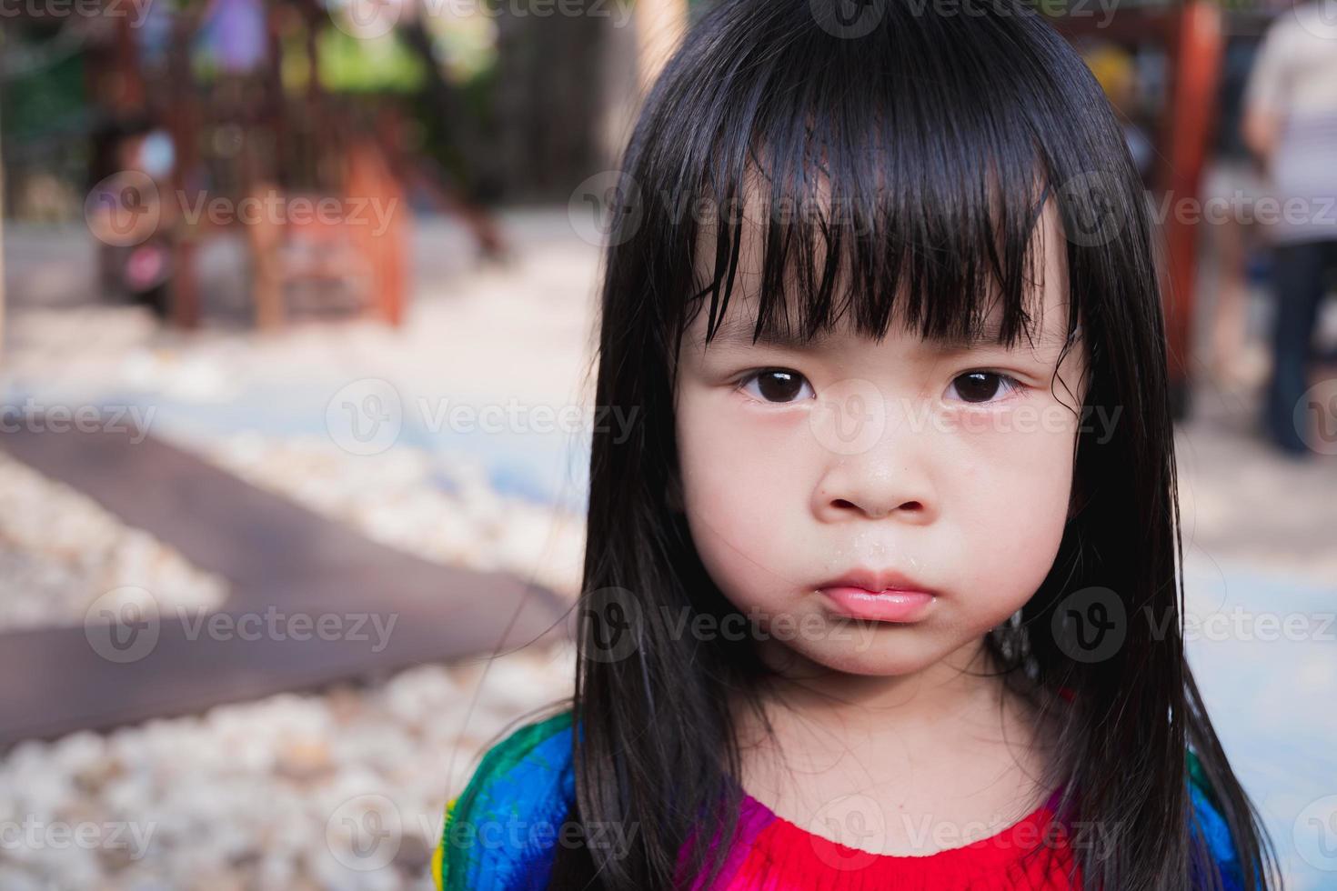 Closeup face child is hot sweat. Upset girl in the playground outside. Kid black haired unhappy when hot weather. Baby wear colorful dress aged 4 years old. In summer or spring time. photo