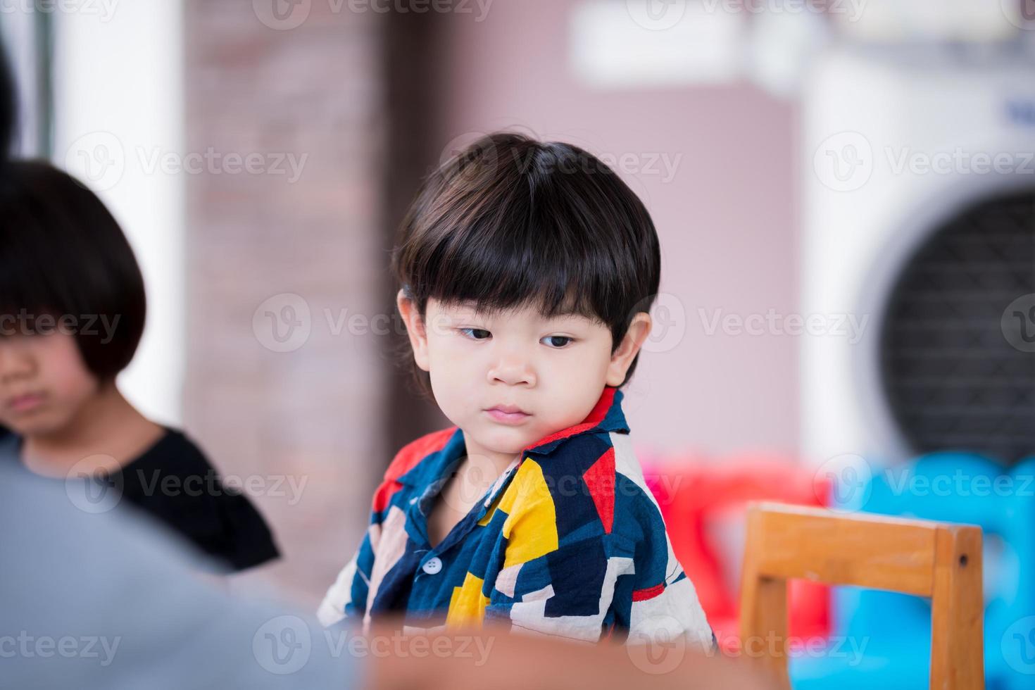 Adorable Asian boy sitting on wooden chair. Baby face sad. Child aged 2 year old, wearing colorful shirt. photo