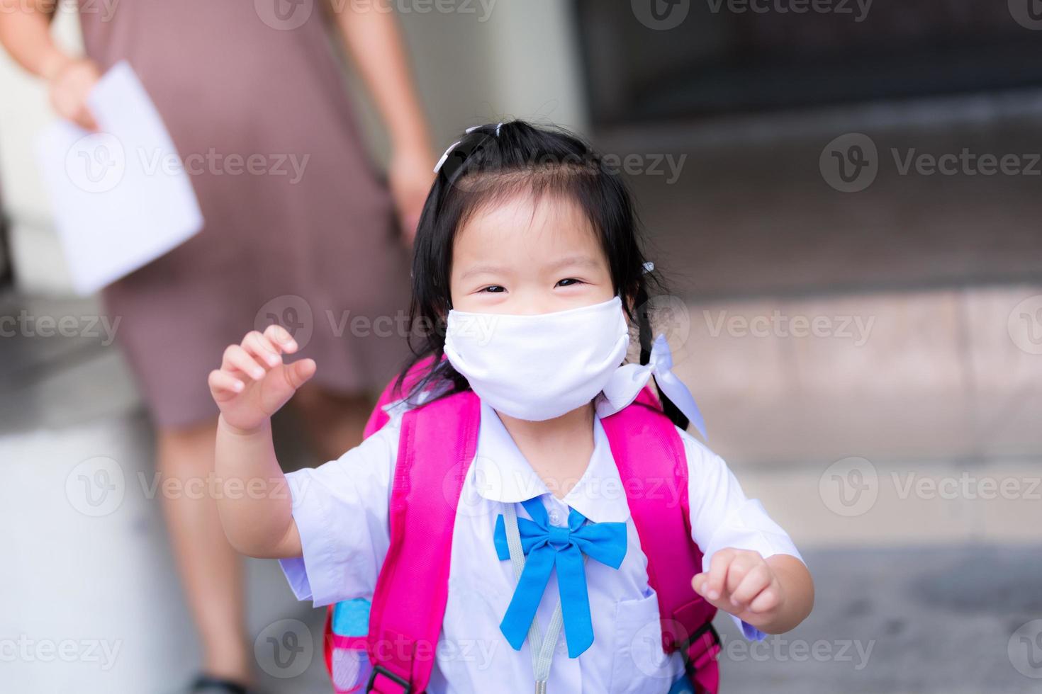 Mother or guardian takes the child back from school. A preschooler ran to see his father carrying a camera with happiness and a smile under a white cloth mask. 3-year-old girl carrying a pink backpack photo