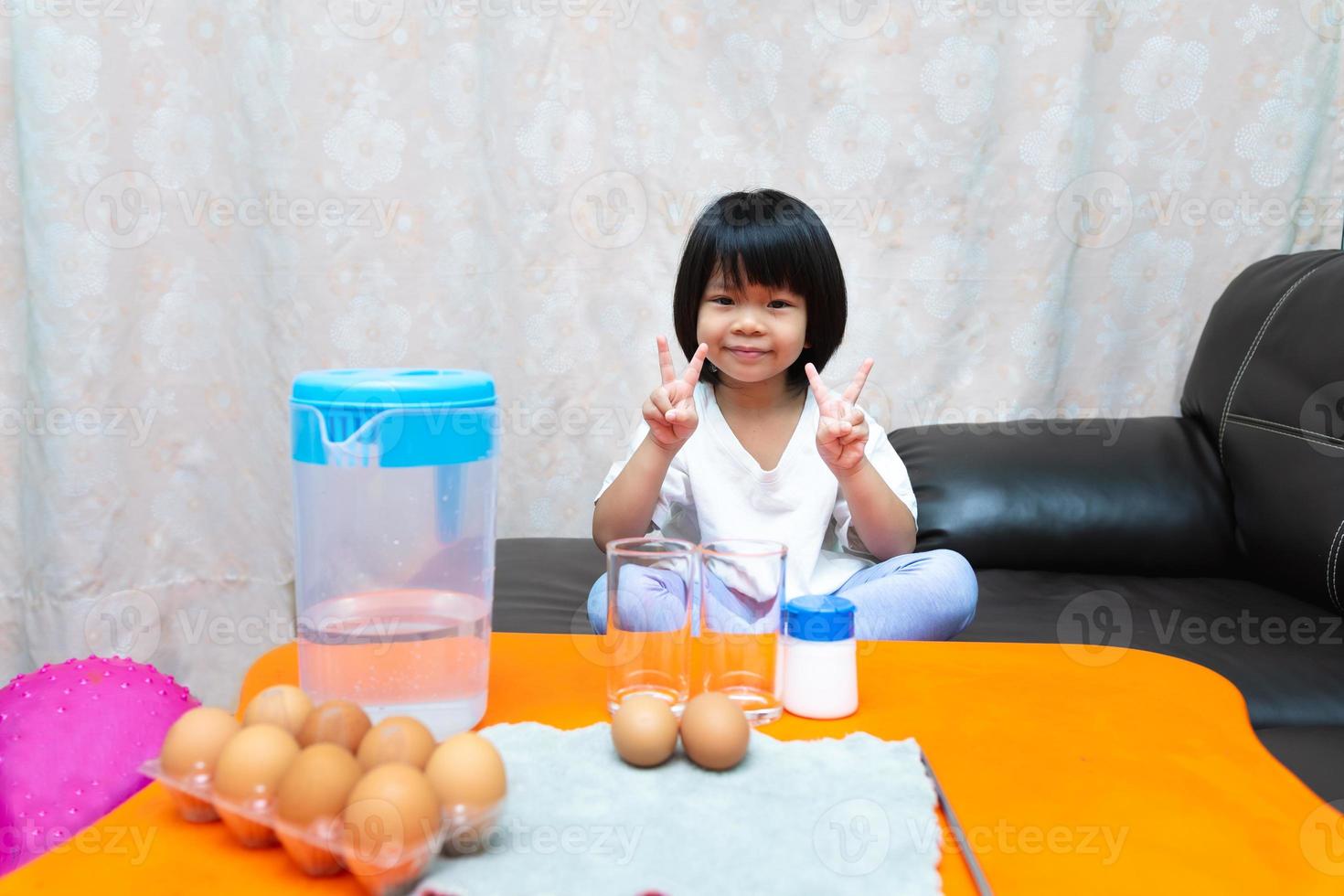 Student sat on sofas waiting to learn experiment. Girl held up two fingers to take picture with sweet smile. Kid will conduct  sunken egg science experiment - the egg floats with water and salt water. photo