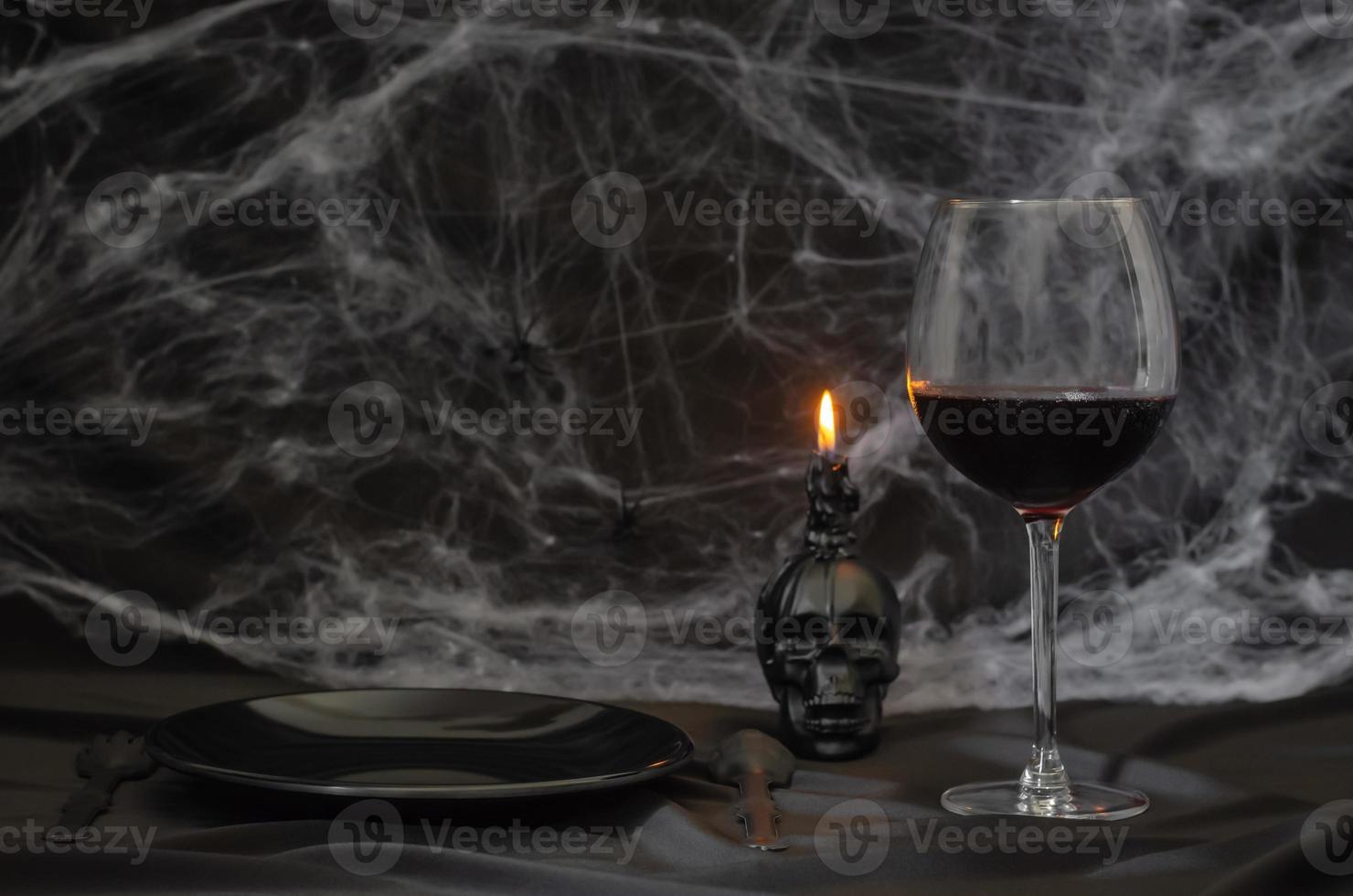 Scary halloween dinner party concept with a glass of red wine, black plate, knife, fork and skull candle with cobweb on black background. photo