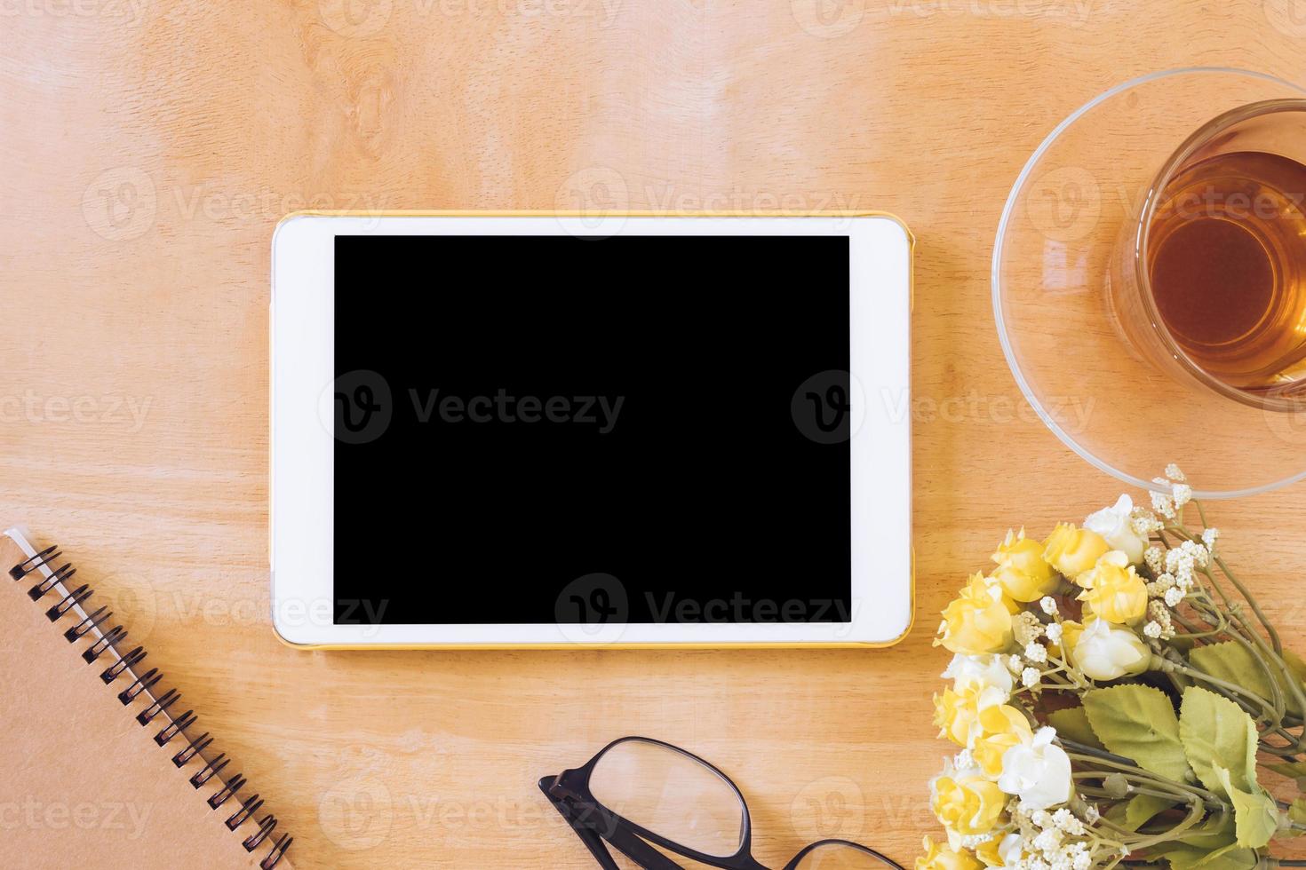 Top view of tablet with tea cup, flower, notebook and glasses on wooden table photo