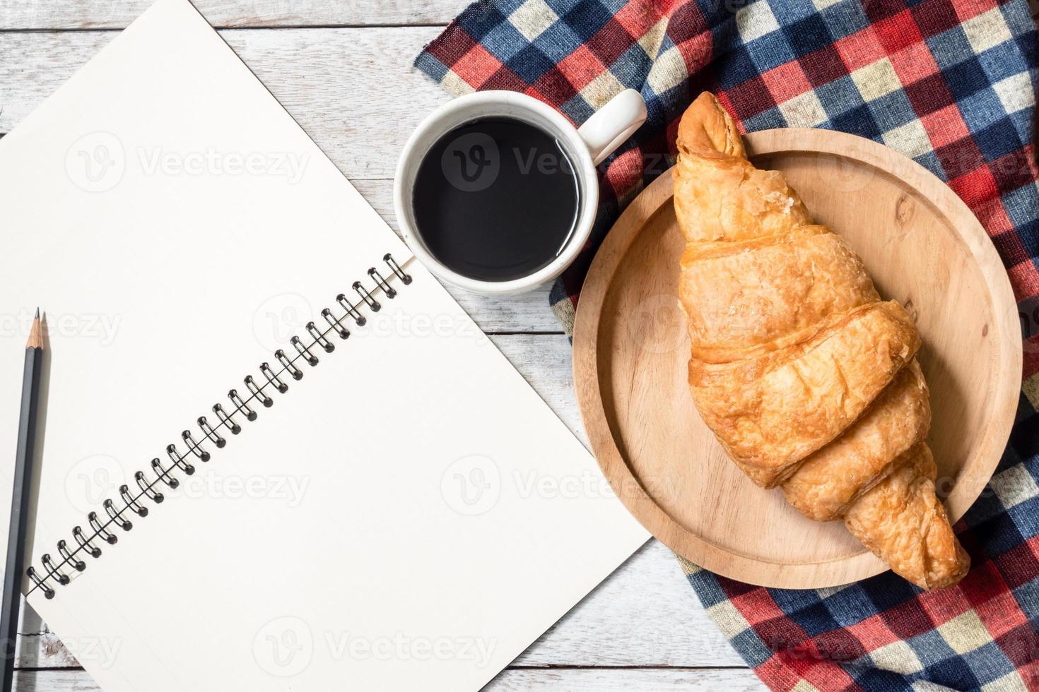 vista superior del cuaderno en blanco con lápiz, croissant y café sobre fondo de mesa de madera. foto